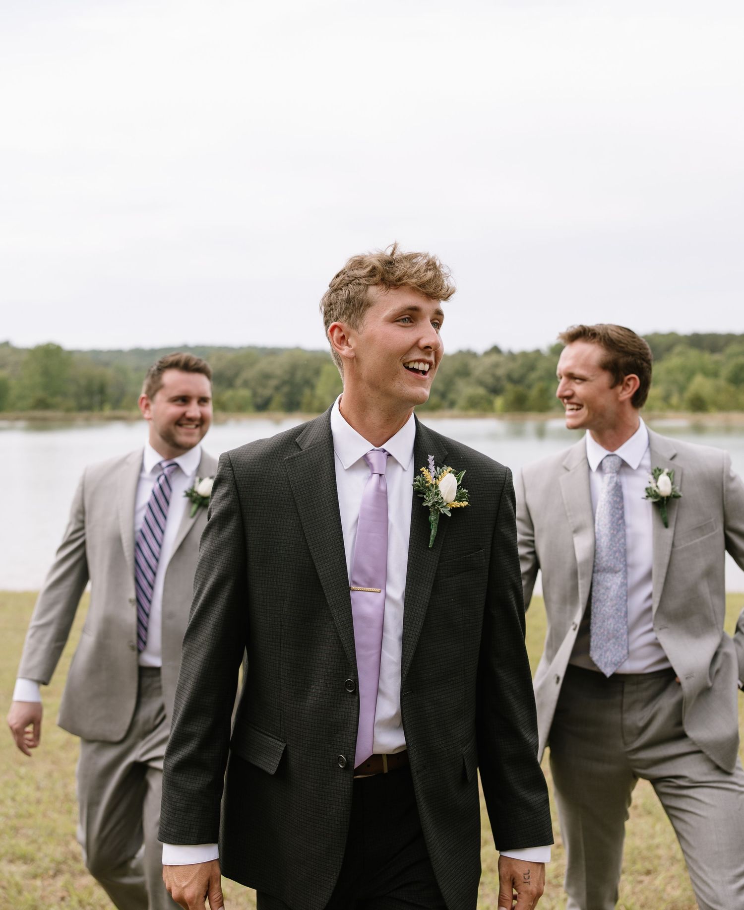 Groom in dark suit with lavender tie and boutonniere walks ahead of groomsmen in gray suits outdoors.