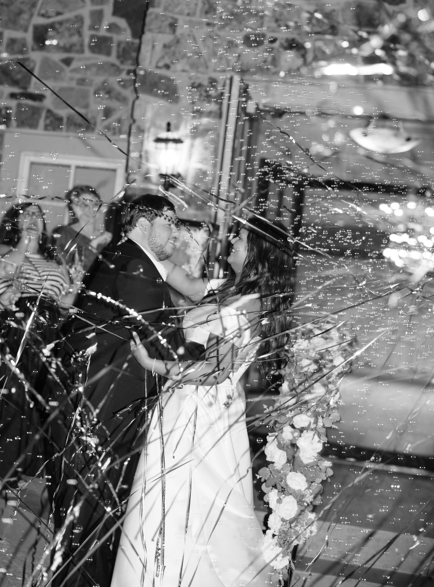 Black and white photo of a bride and groom kissing amid falling confetti streamers at their wedding reception.