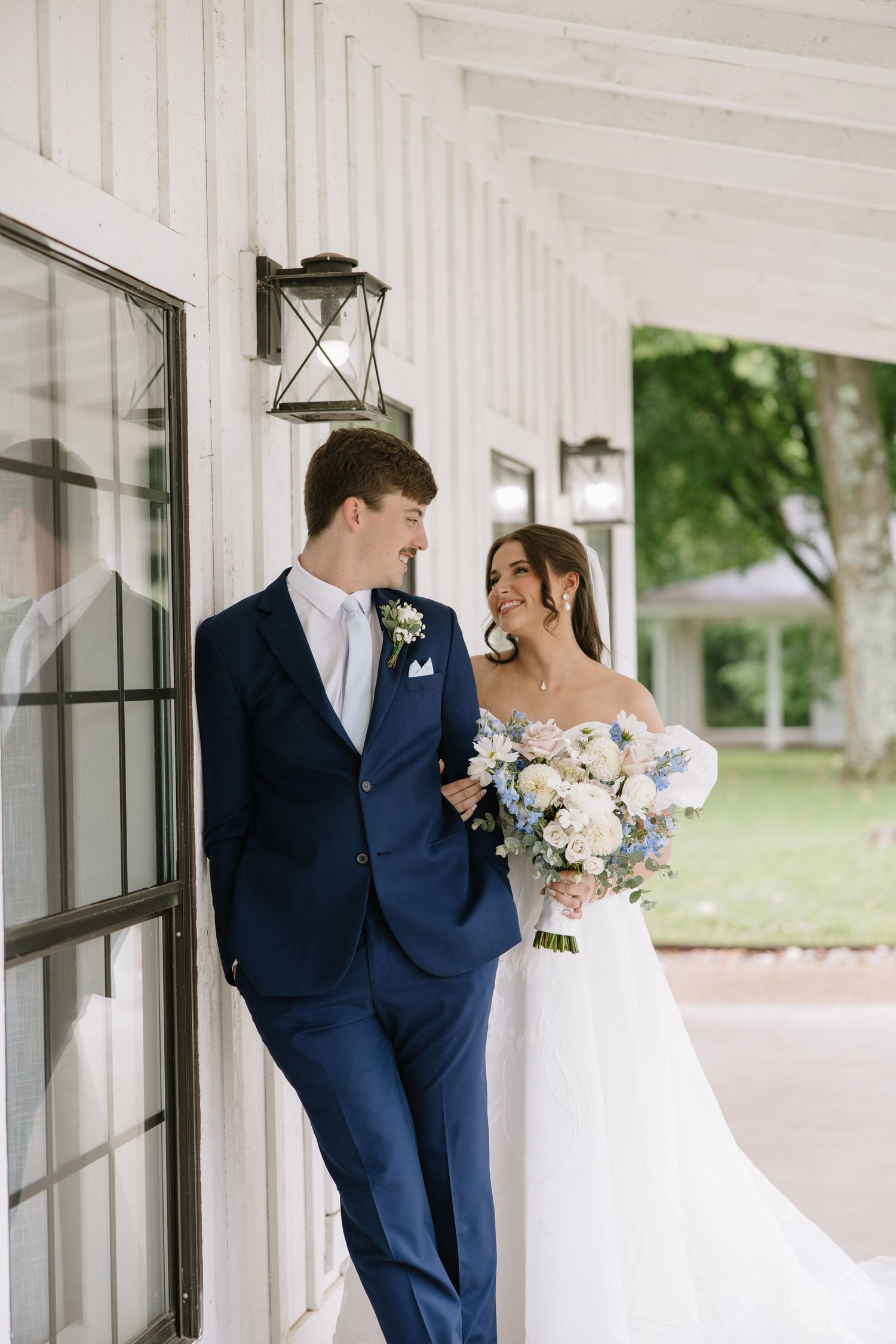 A bride and groom share a romantic moment on a white farmhouse porch, she holds a blue and white bouquet.
