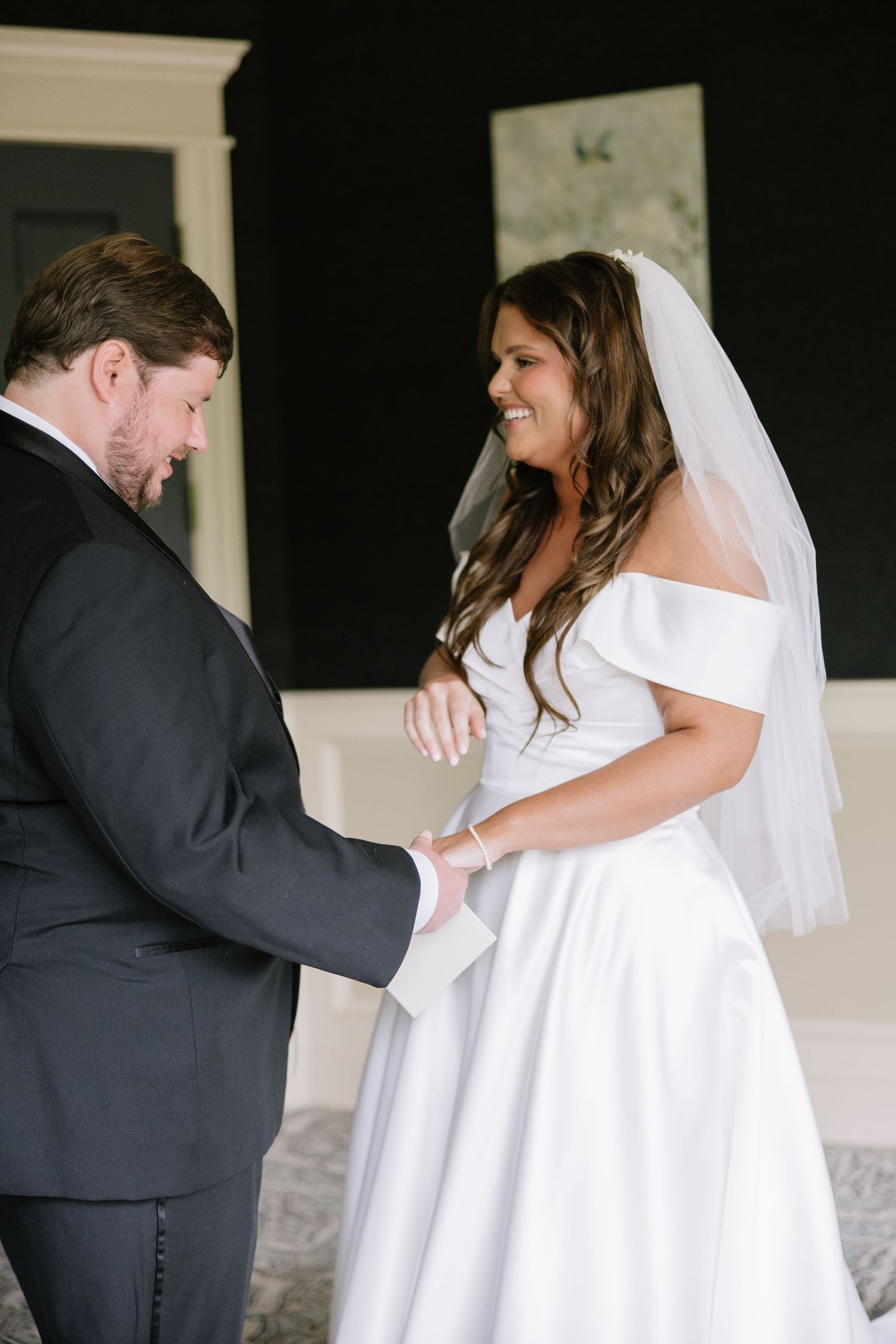 Bride in off-shoulder white gown and veil smiles while holding hands with groom during first look.