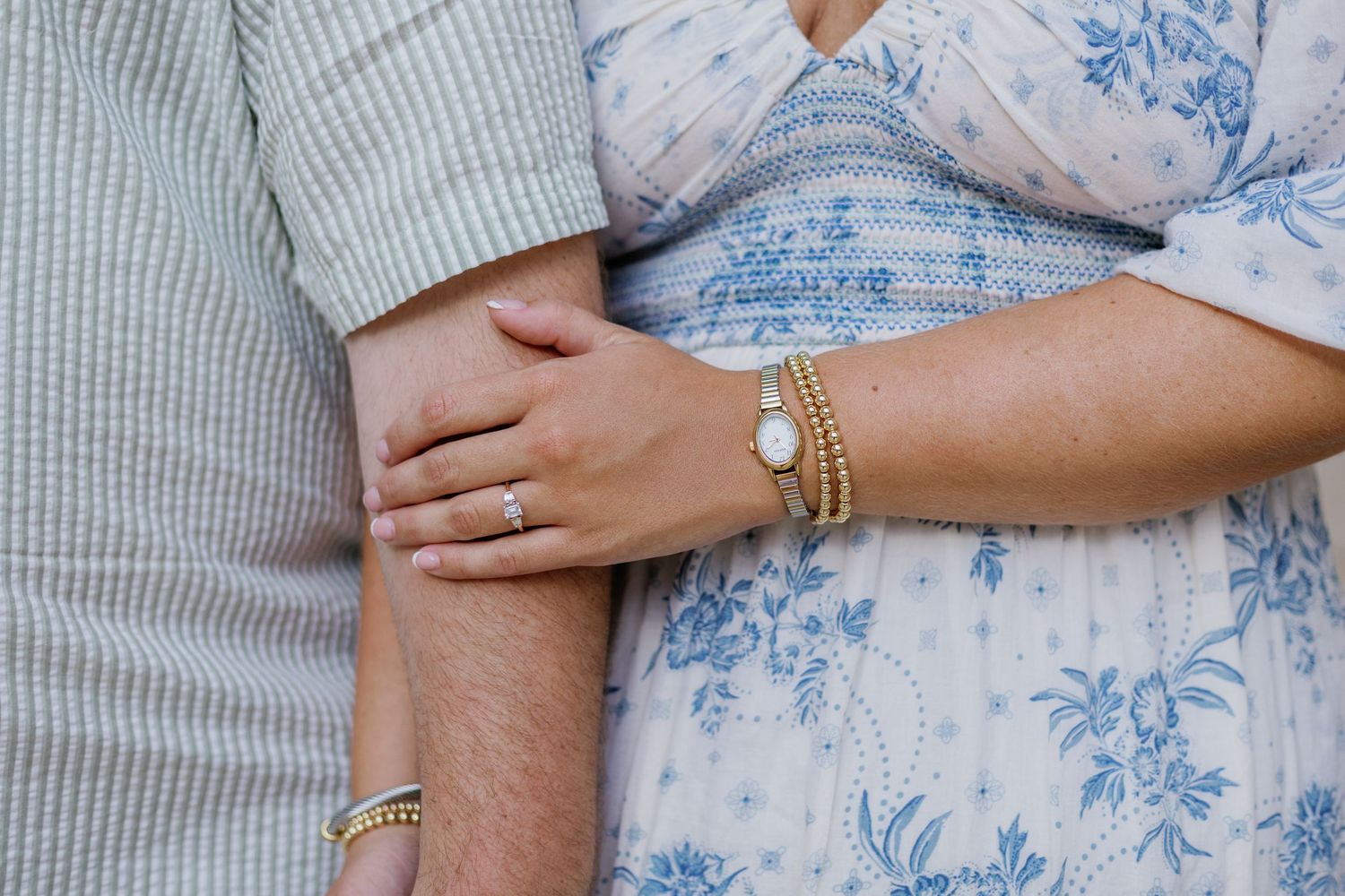 Close-up of couple holding hands, woman wearing engagement ring, gold bracelet, and blue floral dress beside man in striped shirt.