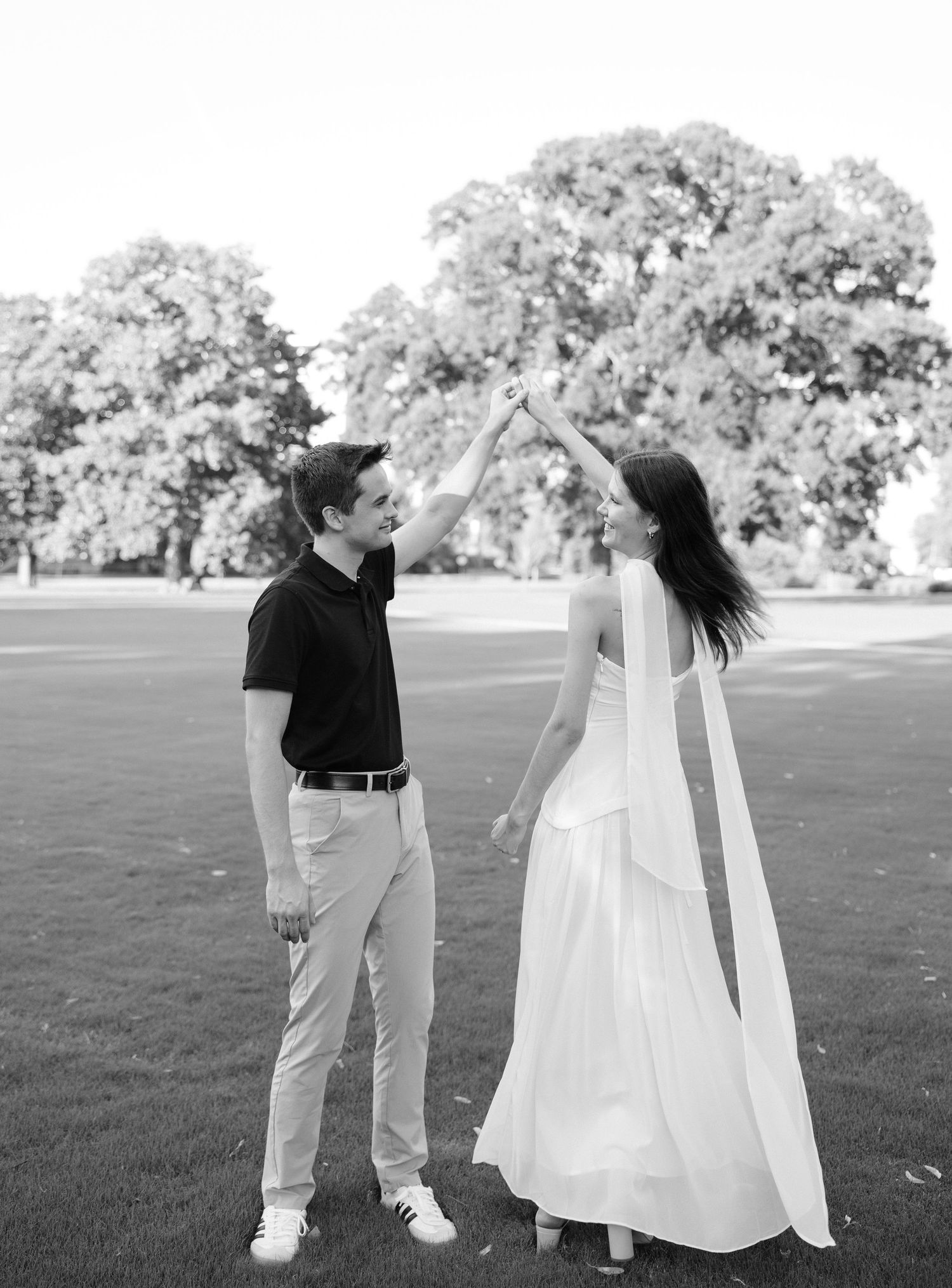 A couple dances outdoors in black and white, bride in flowing white gown and veil, groom in black shirt, trees behind.