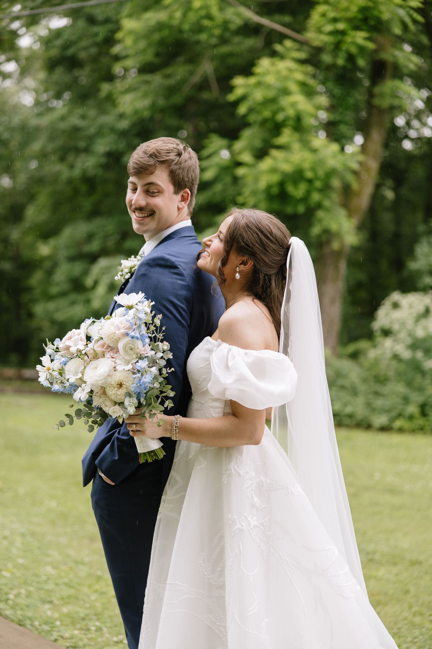 Bride and groom pose back-to-back outdoors, she holds blue and white bouquet, wearing white gown and veil.