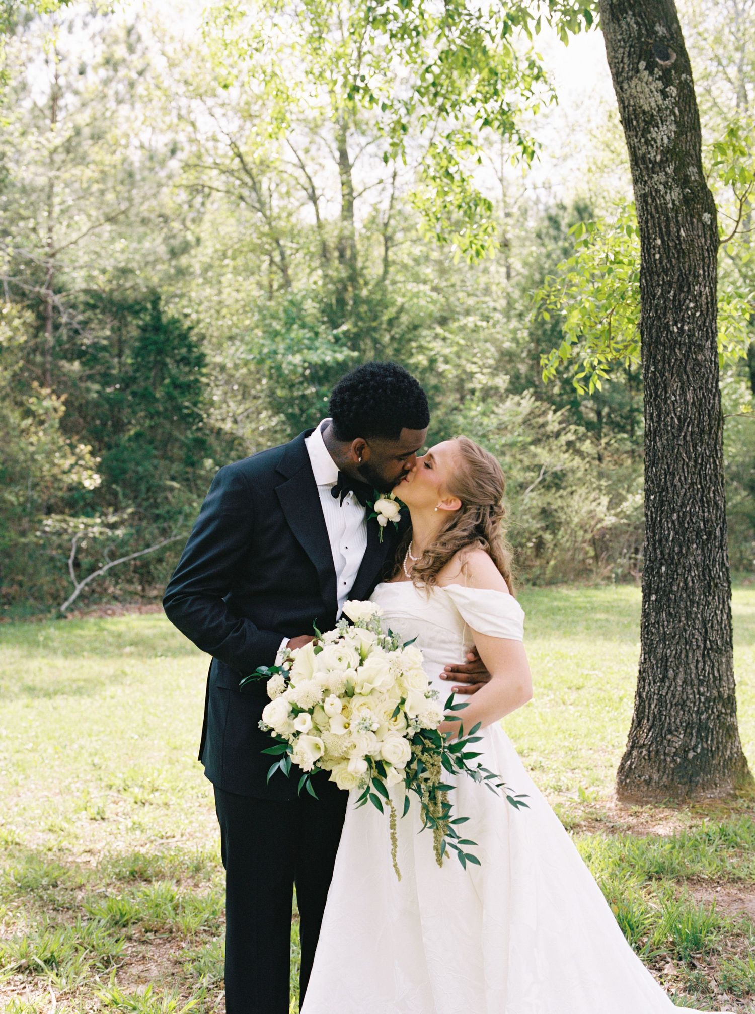 Bride and groom share a kiss outdoors under trees, bride holding white floral bouquet.