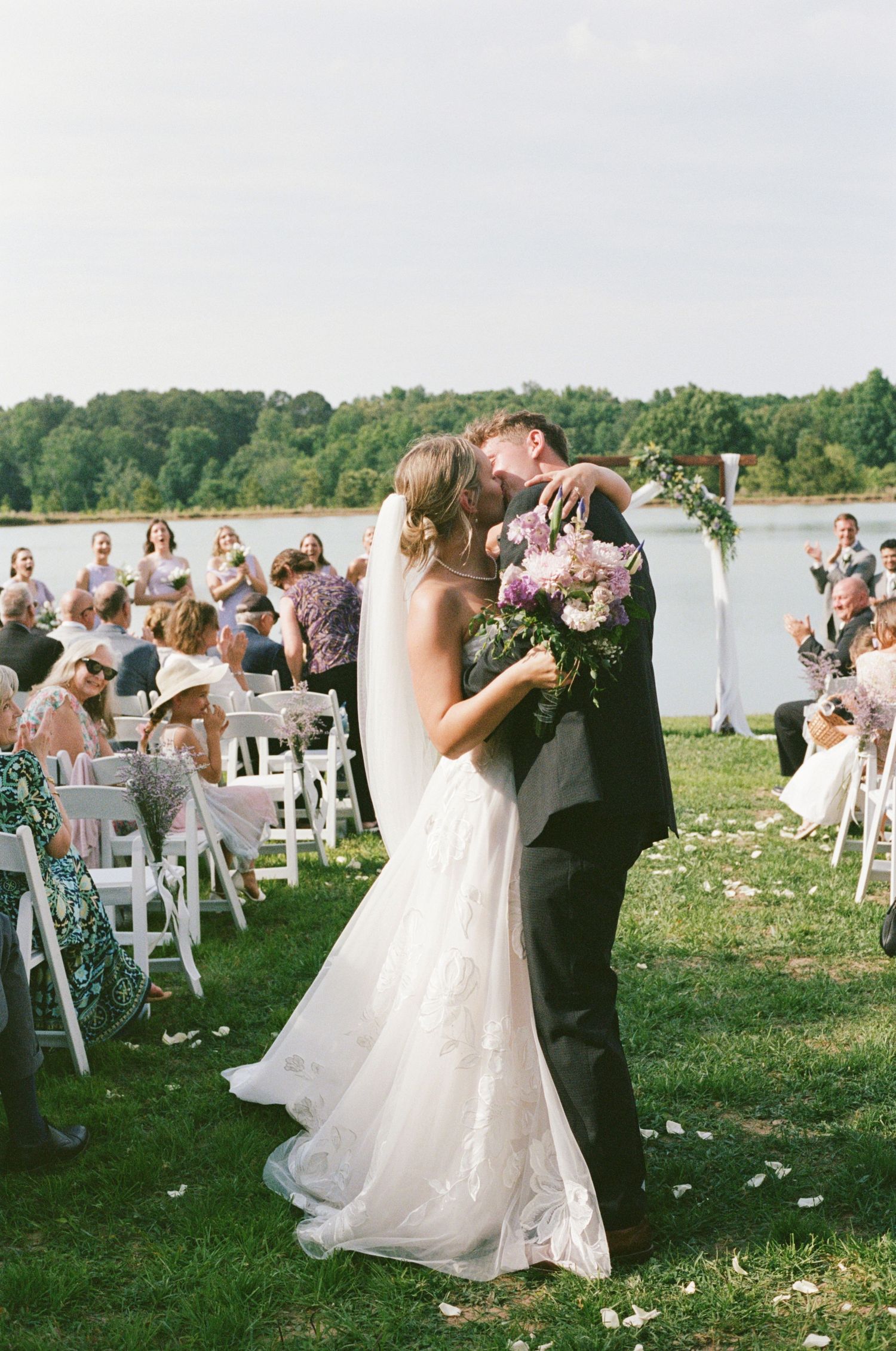 Bride and groom share first kiss at outdoor lakeside wedding ceremony with white chairs and floral bouquet.