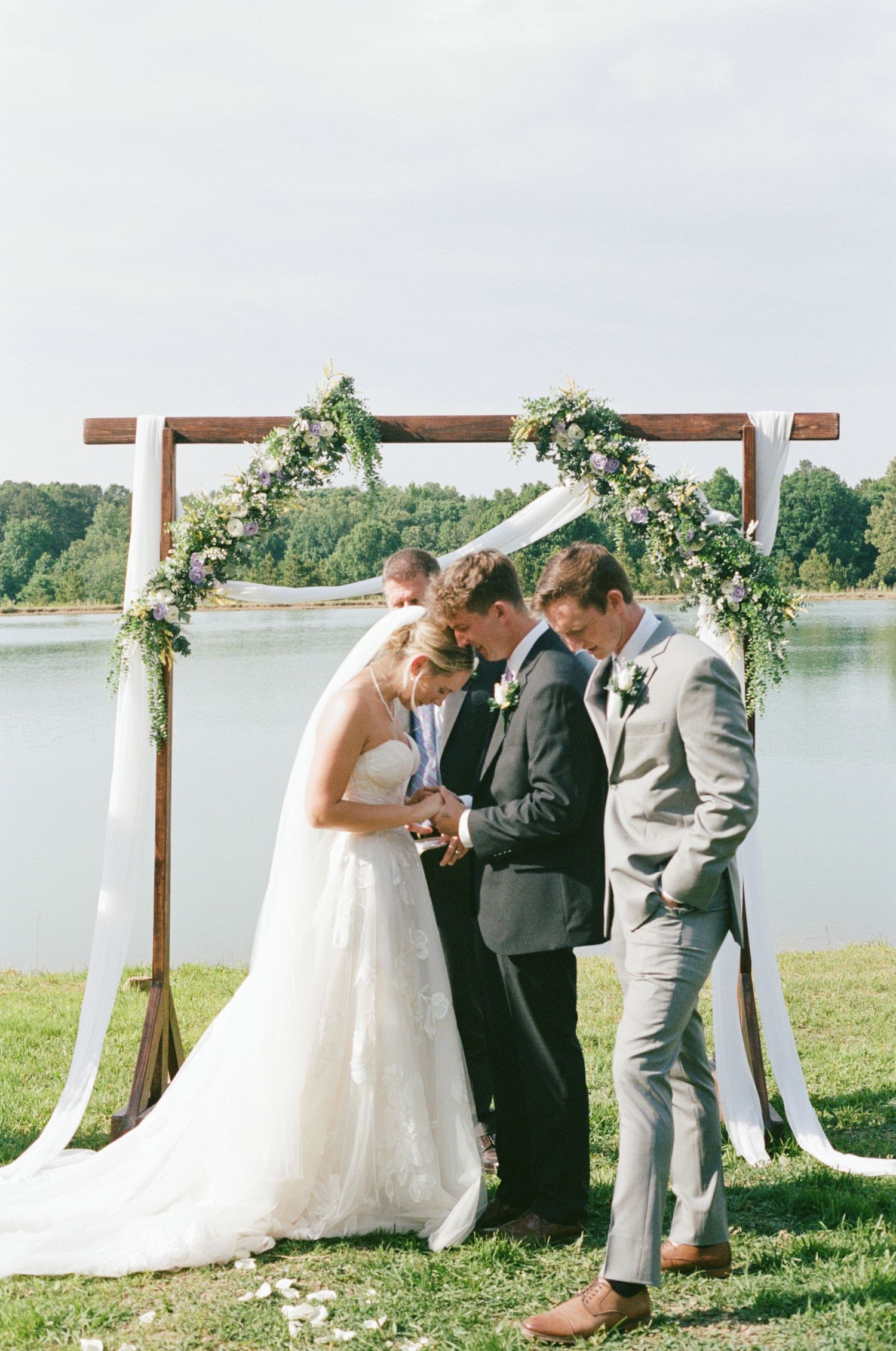 Bride and groom exchange vows under a rustic wooden arch adorned with flowers by a lakeside on a sunny wedding day.