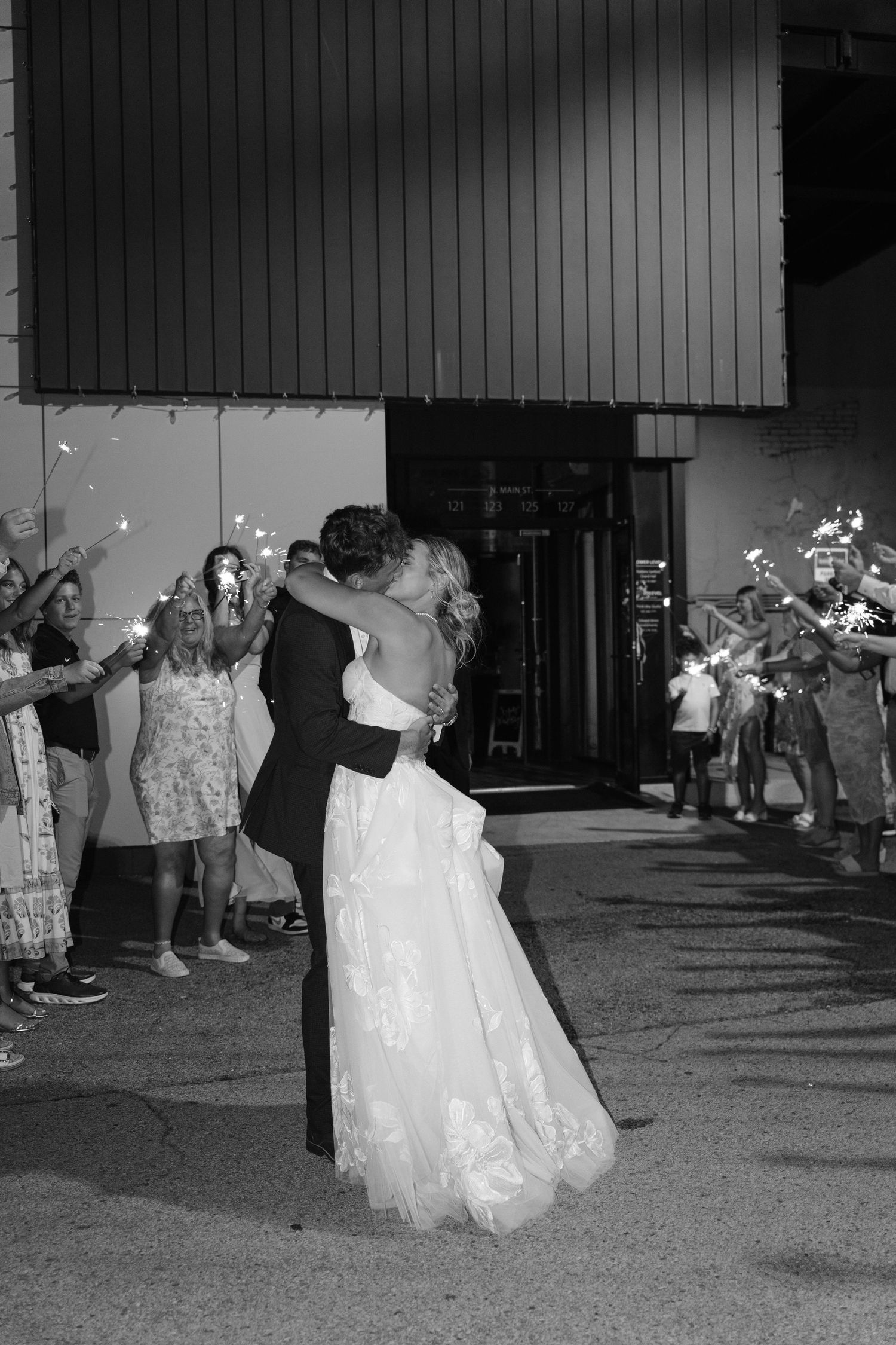 Newlyweds sharing a kiss during sparkler send-off with cheering guests in black and white.