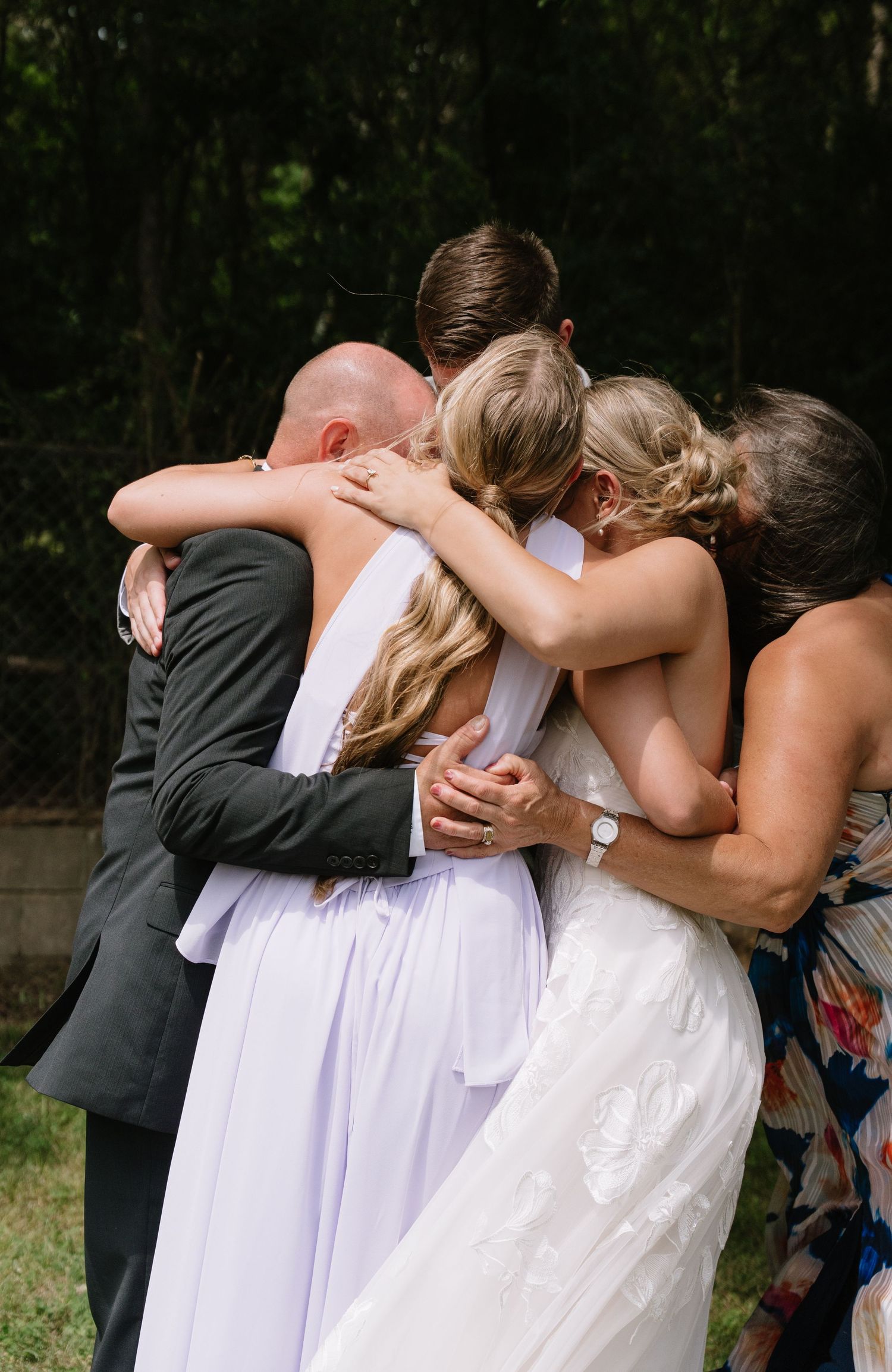 Wedding group hug with bride in white gown and veil embracing family members outdoors.