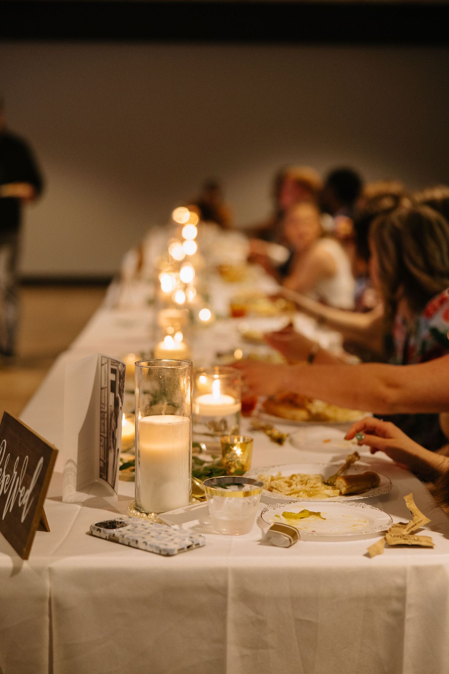 Elegant candlelit dinner table with white linens and guests dining in a warmly lit venue.