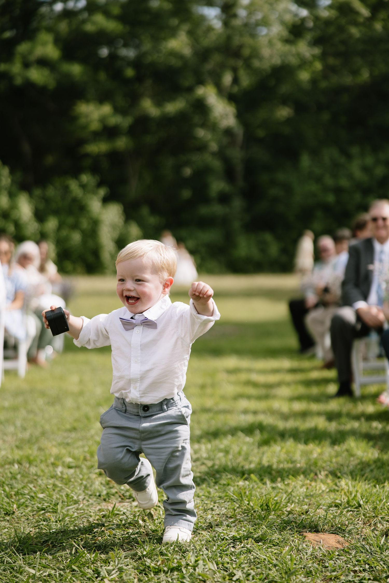 Joyful toddler ring bearer in white shirt and gray pants runs down outdoor wedding aisle holding small gift.