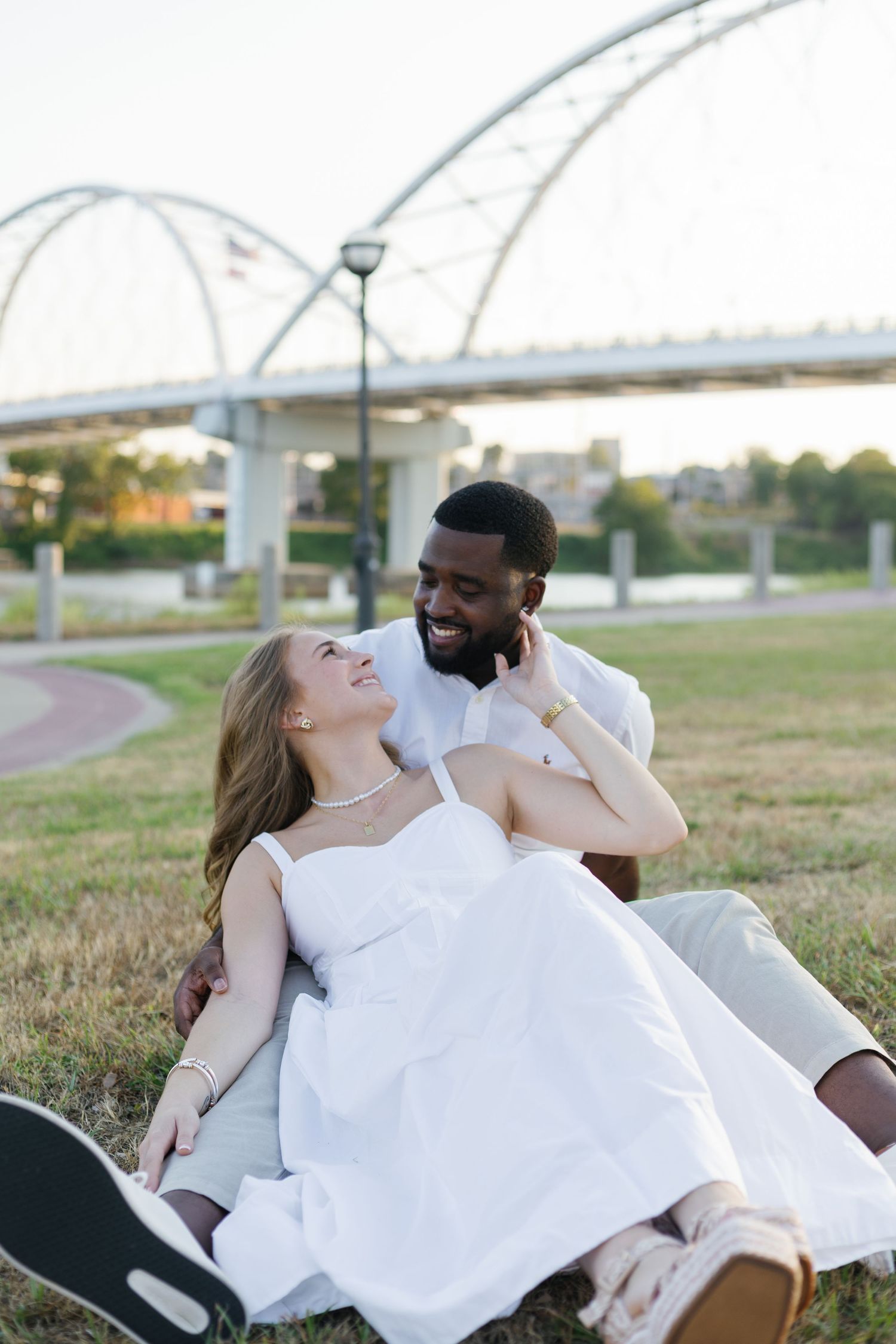 A happy interracial couple sits on grass in a park, smiling at each other, with a bridge and arch structures in the background.