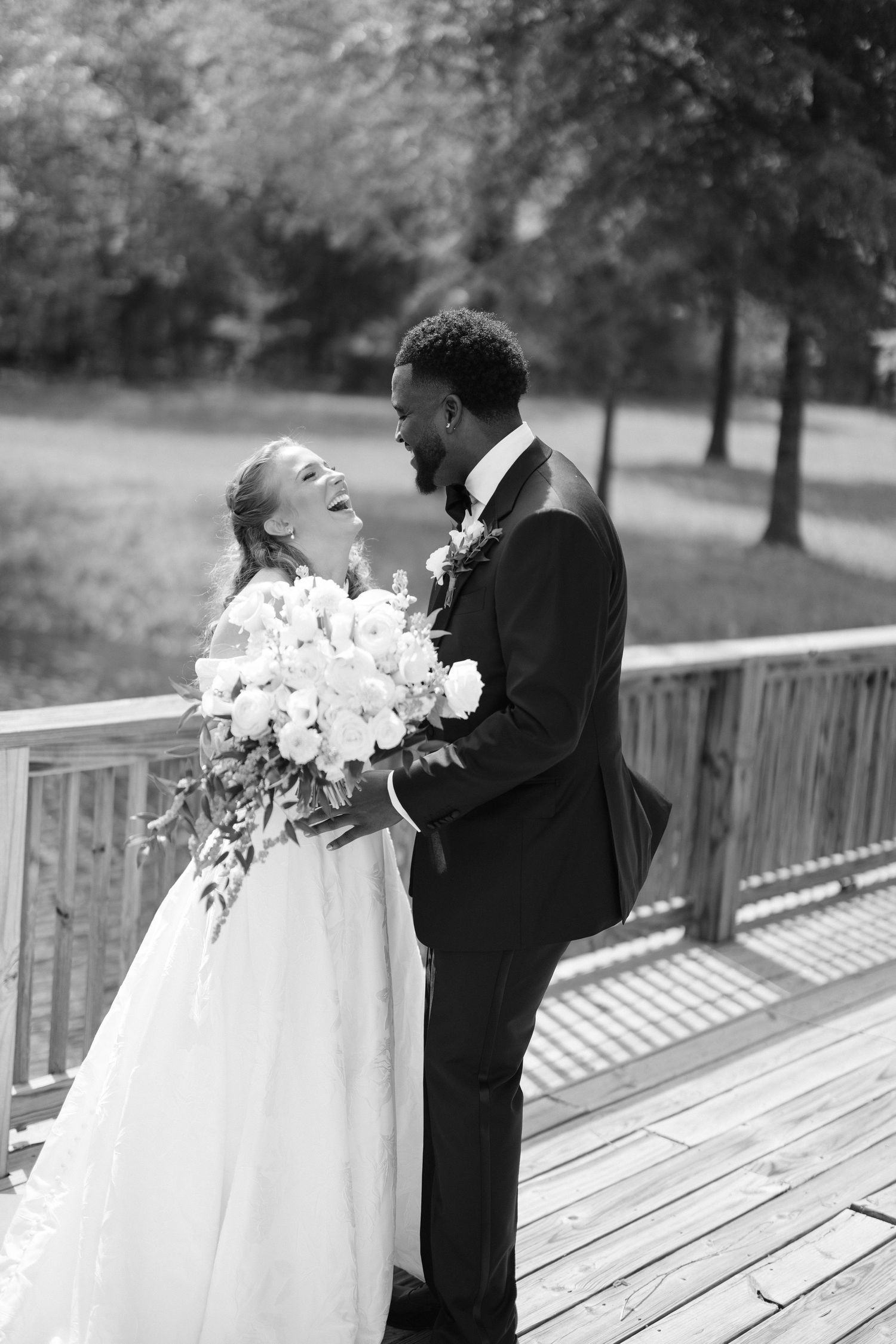 Black and white photo of interracial bride and groom laughing together on a wooden bridge holding white bouquet.