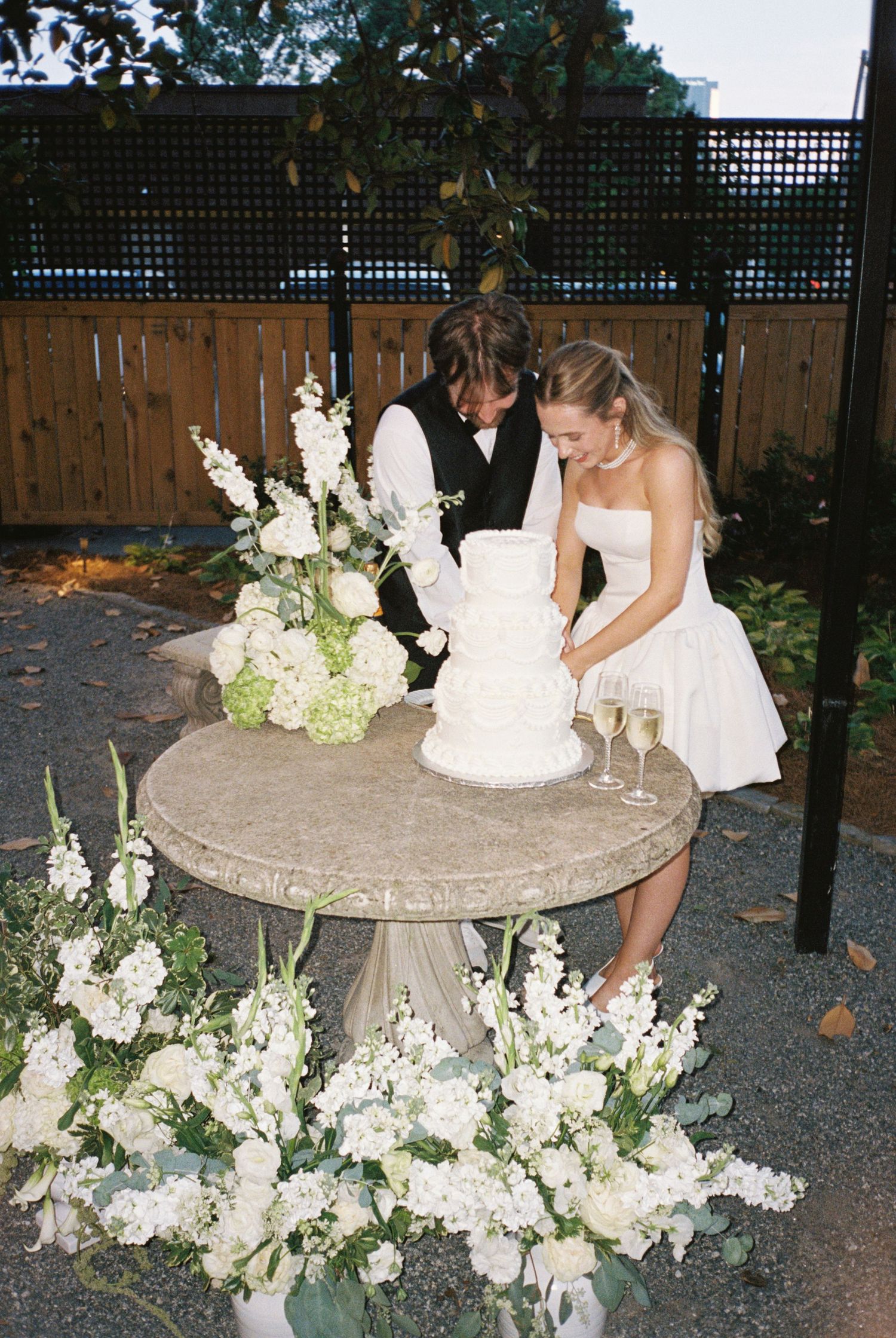 Bride and groom cut a three-tier white wedding cake surrounded by white floral arrangements.