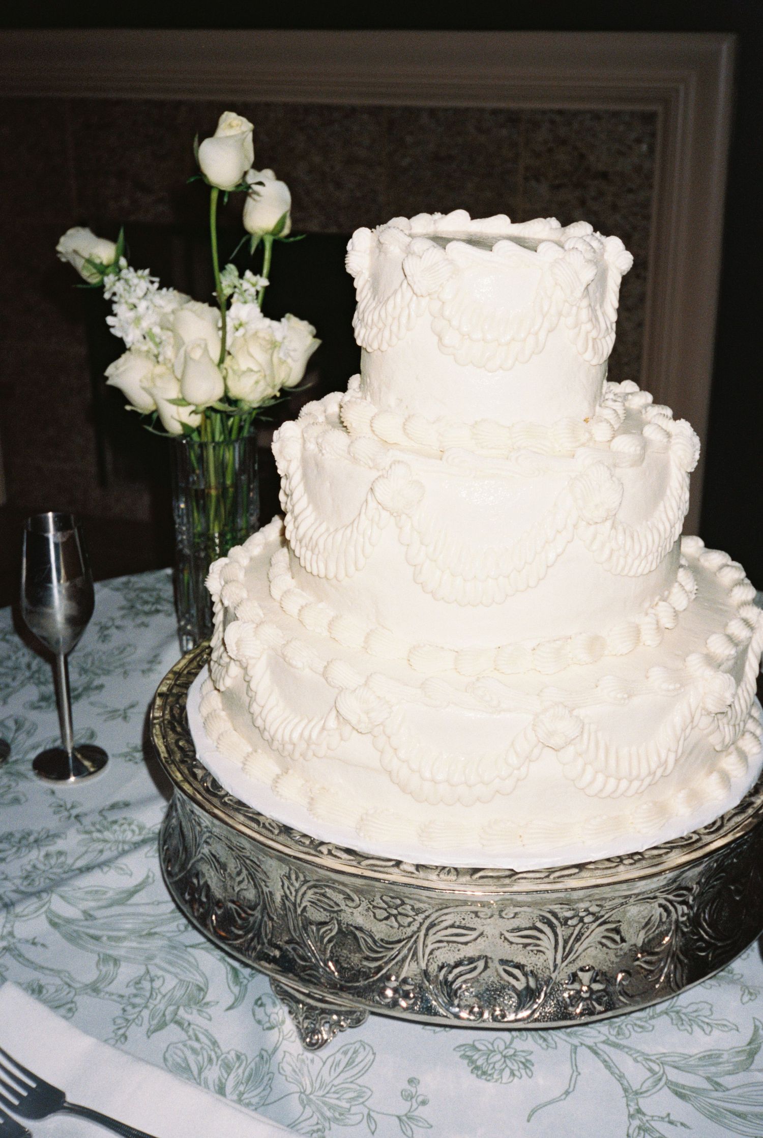 Elegant three-tier white wedding cake with intricate piped frosting details on a silver stand with white roses.