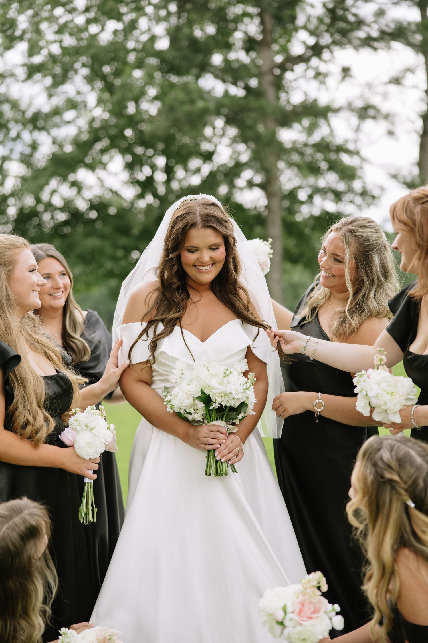 Bride in white off-shoulder gown holding bouquet surrounded by bridesmaids in black dresses outdoors.