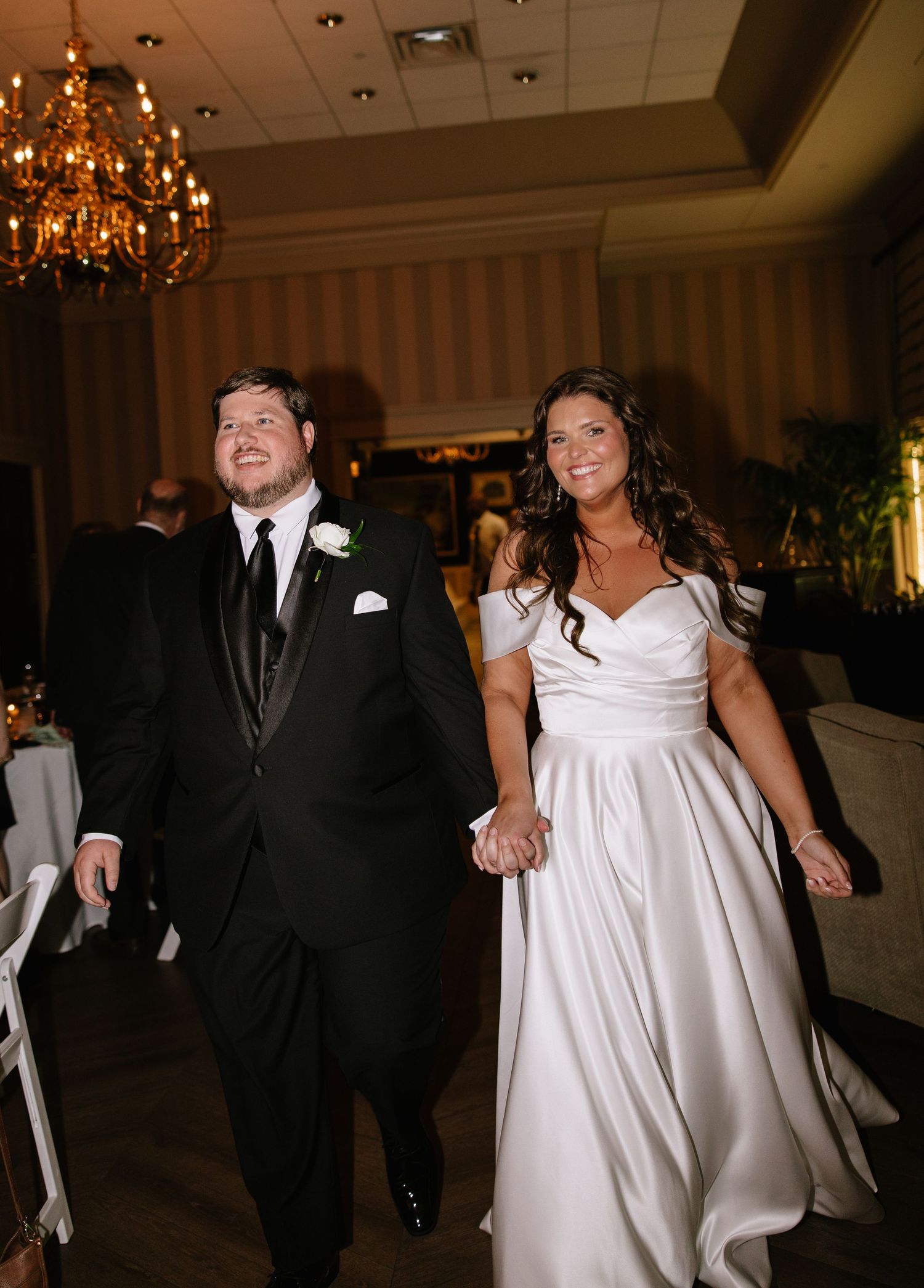 Newlyweds enter wedding reception hand-in-hand in elegant ballroom with chandelier lighting.