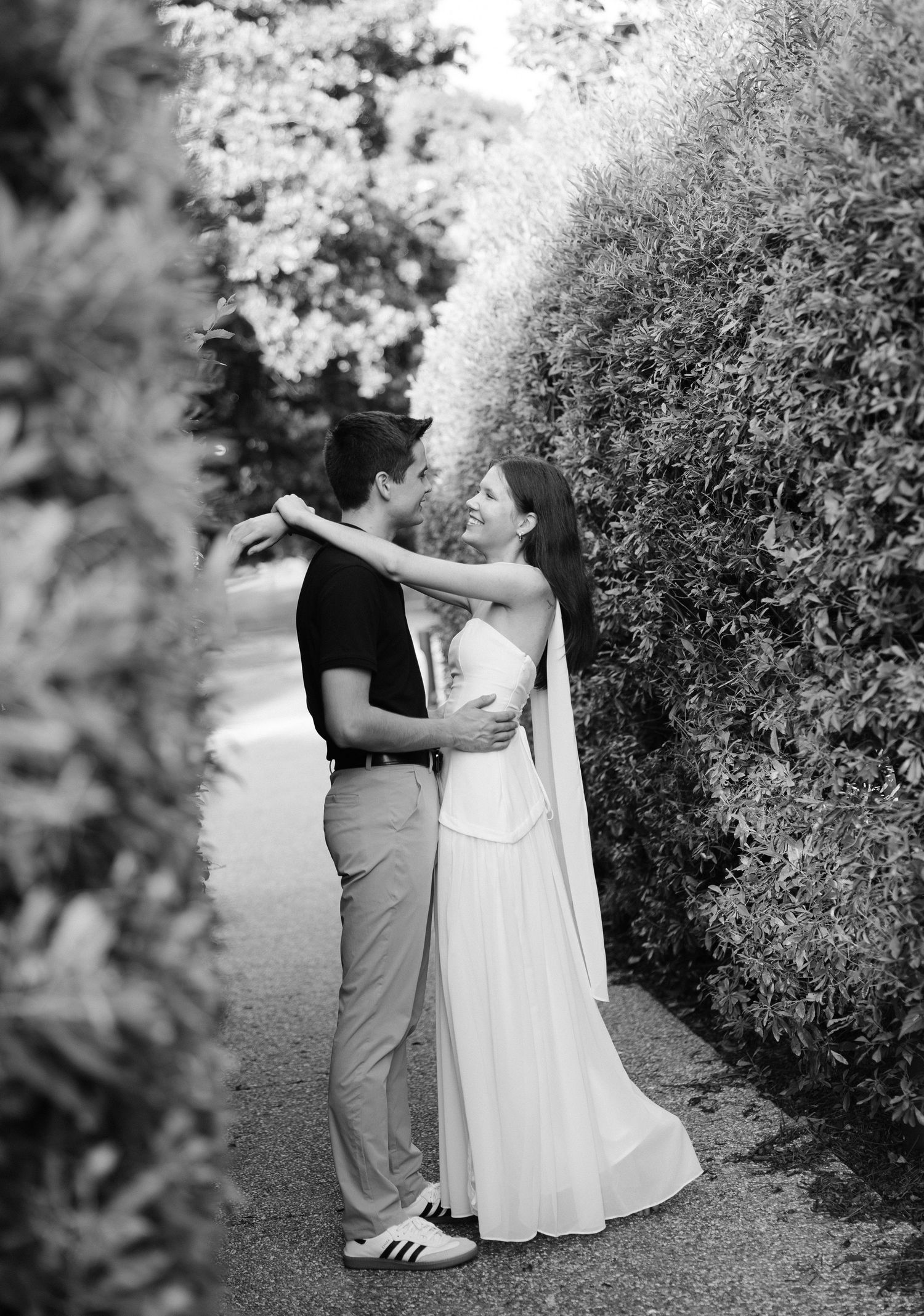 Black and white photo of couple embracing and kissing near a lush hedge during engagement shoot.