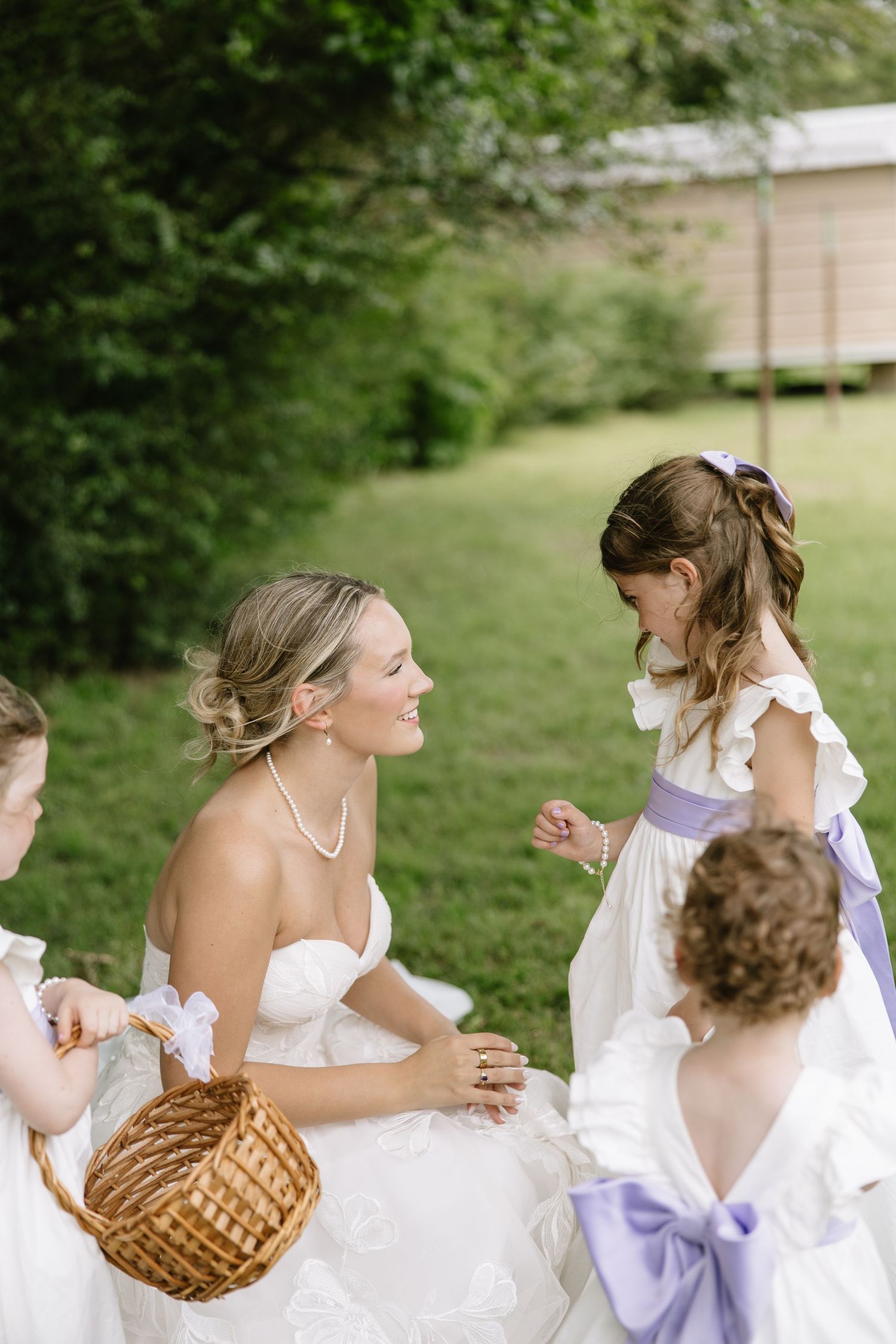 Bride in white strapless gown smiles at flower girls in purple sashes during outdoor wedding ceremony.