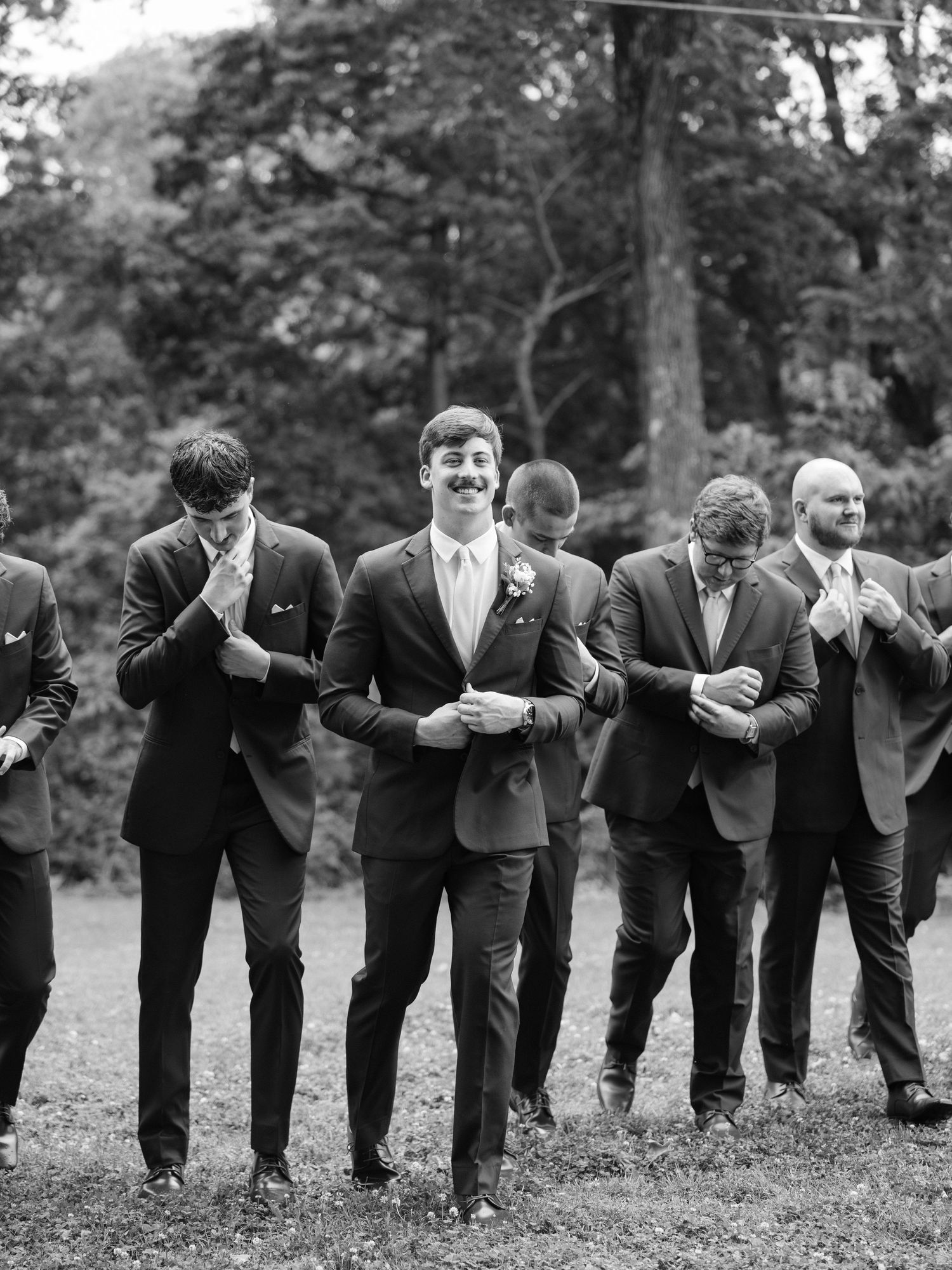 Black and white photo of groom and groomsmen in suits adjusting cufflinks outdoors at a wedding.