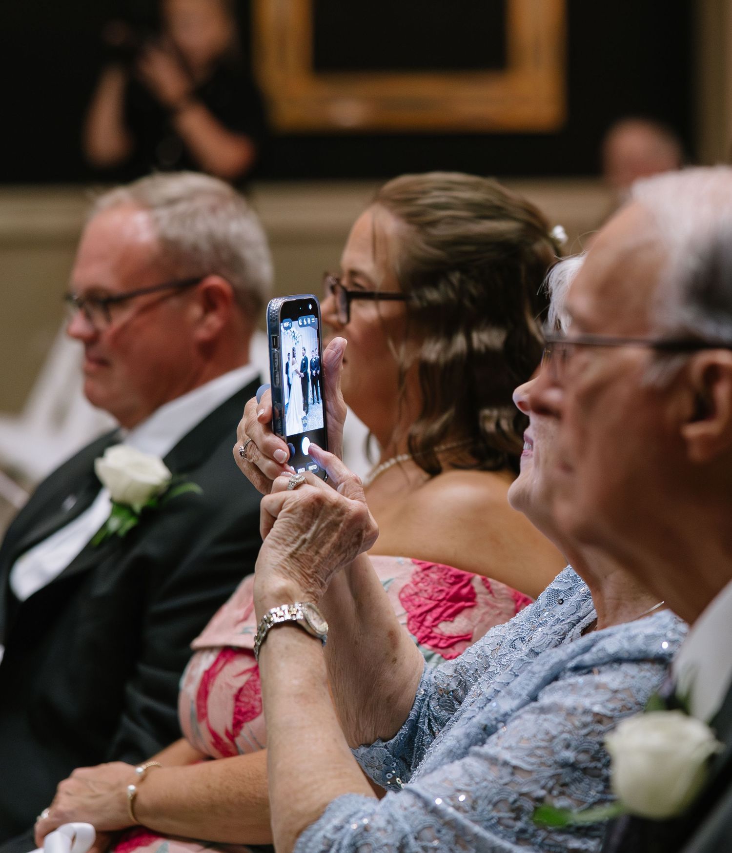 Wedding guests seated in elegant venue, woman in pink dress photographs ceremony on smartphone.