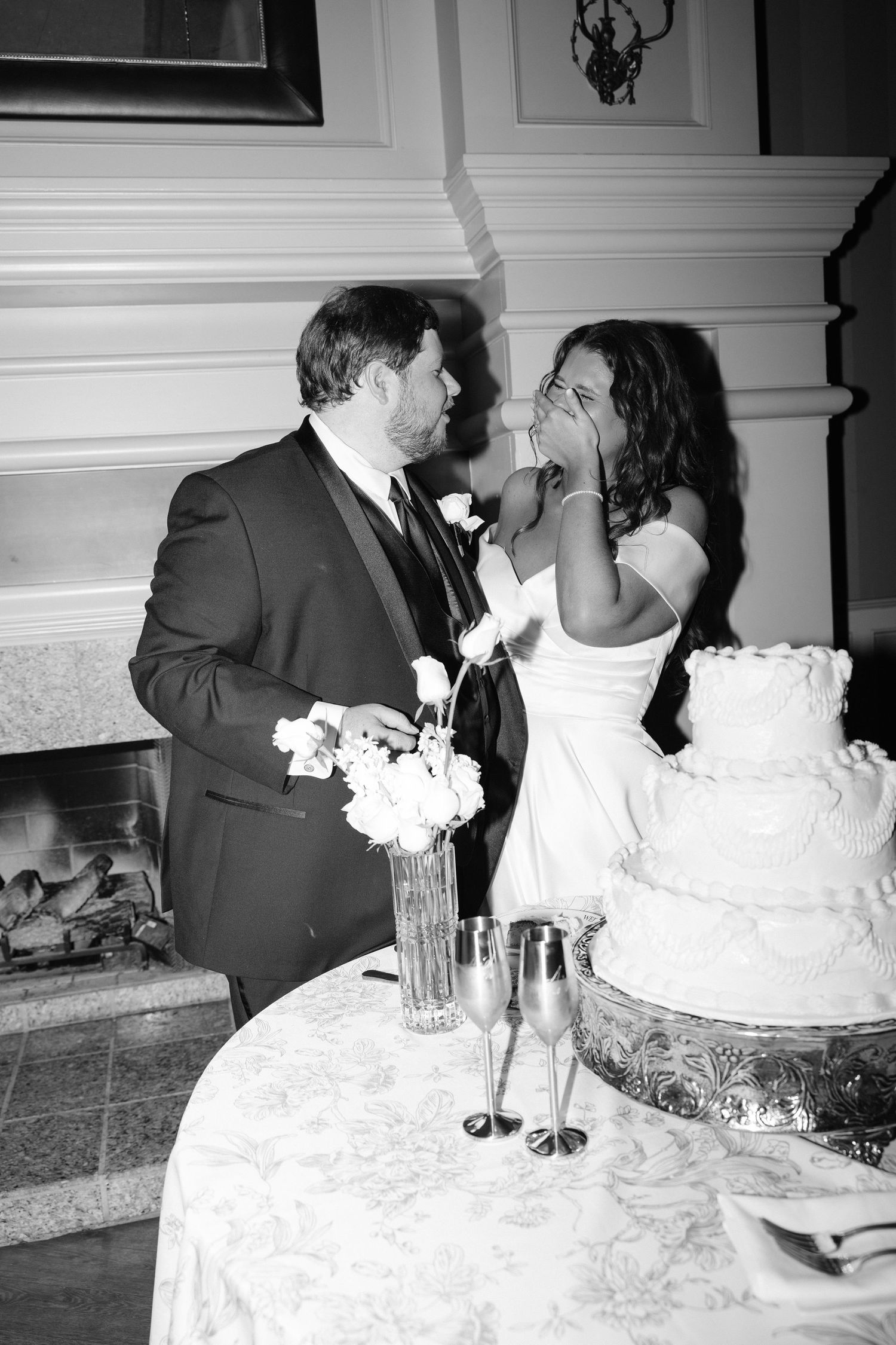 Black and white photo of a bride and groom sharing a moment by their wedding cake near a fireplace.
