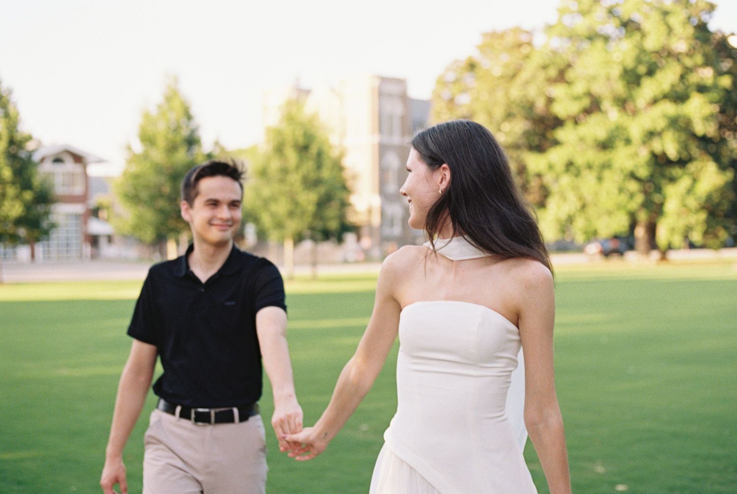 Couple holding hands on green lawn, woman in white dress and man in black polo shirt, trees in background.