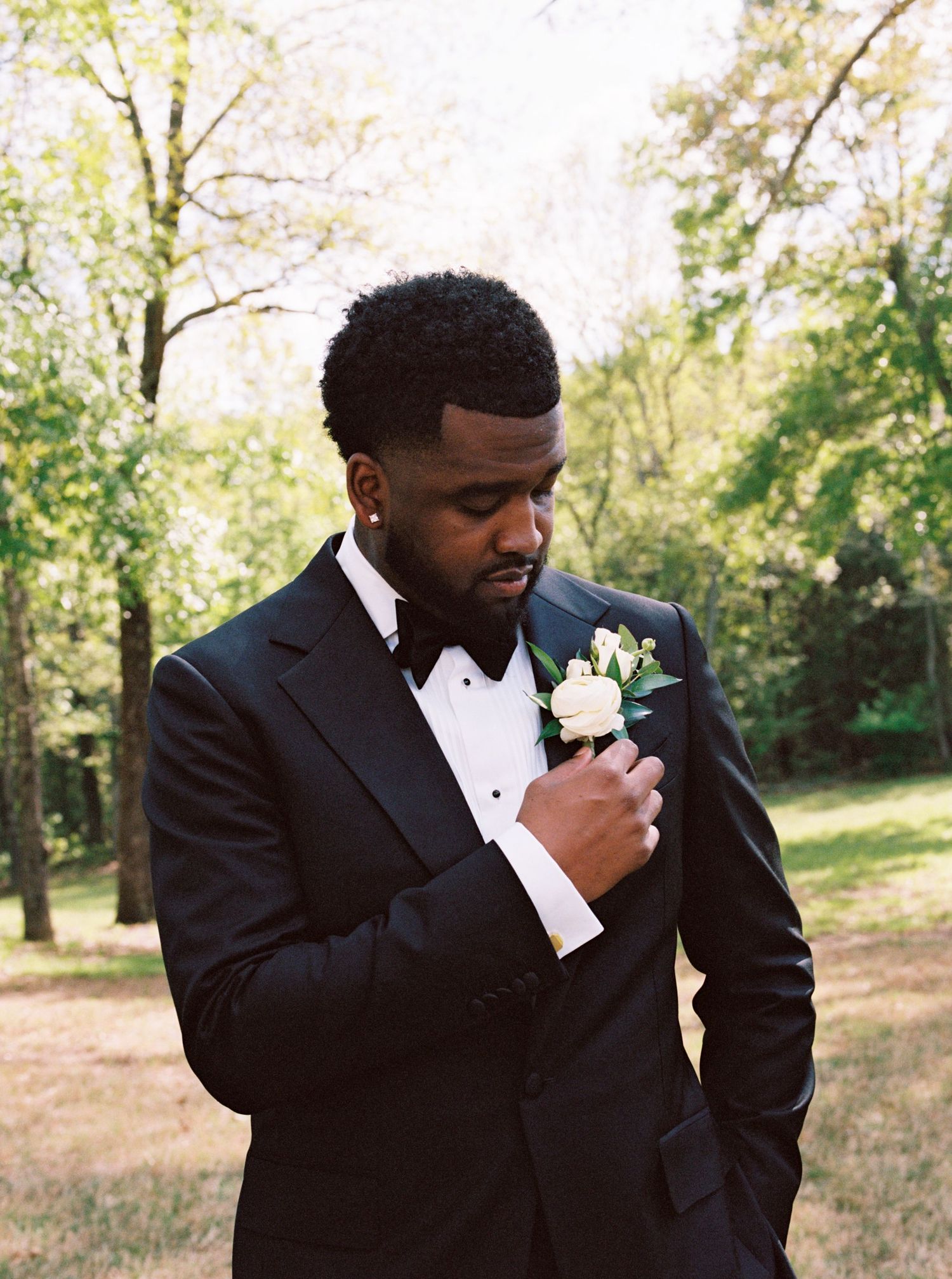 Dapper groom in black tuxedo adjusting white rose boutonniere outdoors among trees.