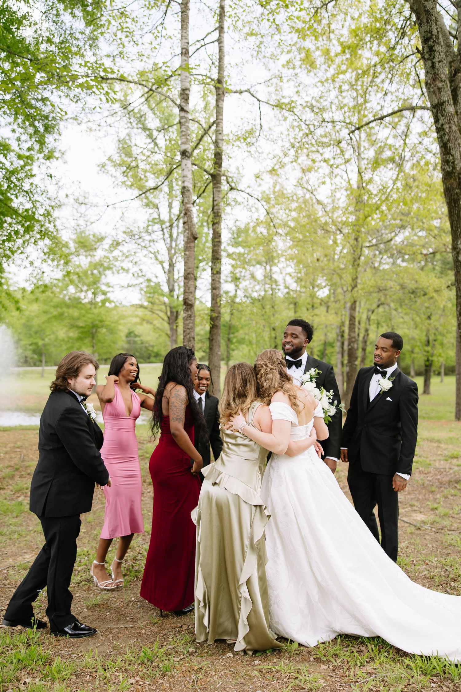 Wedding party in formal attire celebrating with the interracial bride and groom in a lush green outdoor setting.