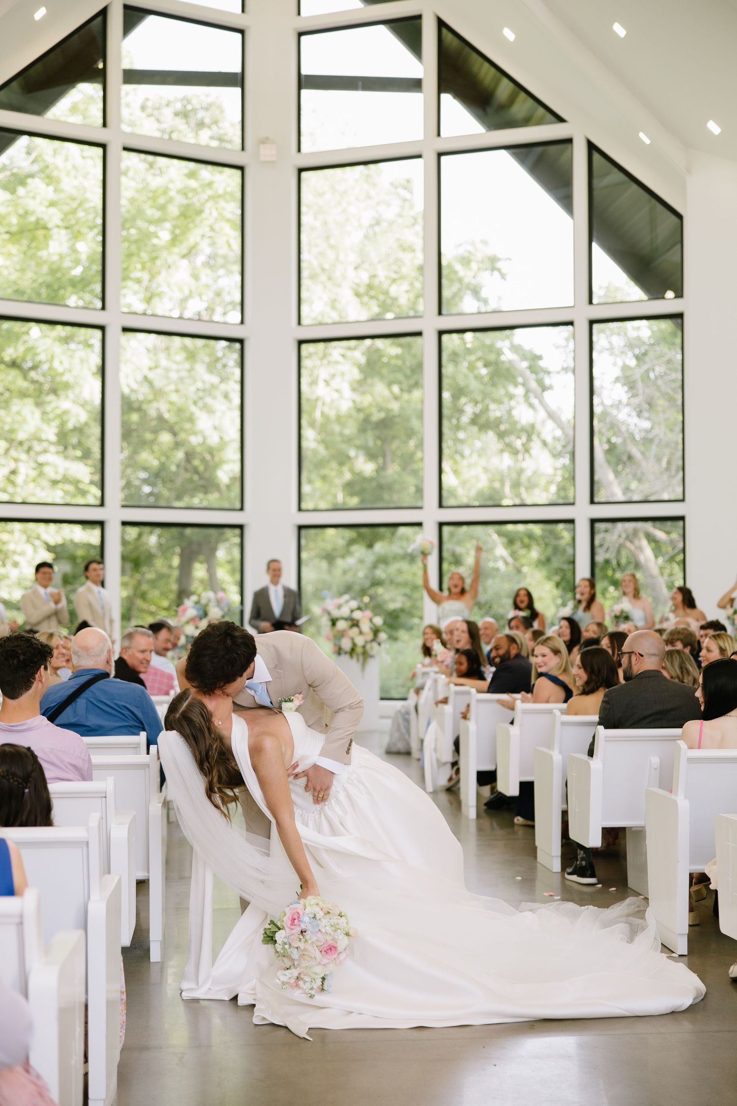 Bride and groom share first kiss at altar in elegant white chapel with floor-to-ceiling windows and cheering guests.