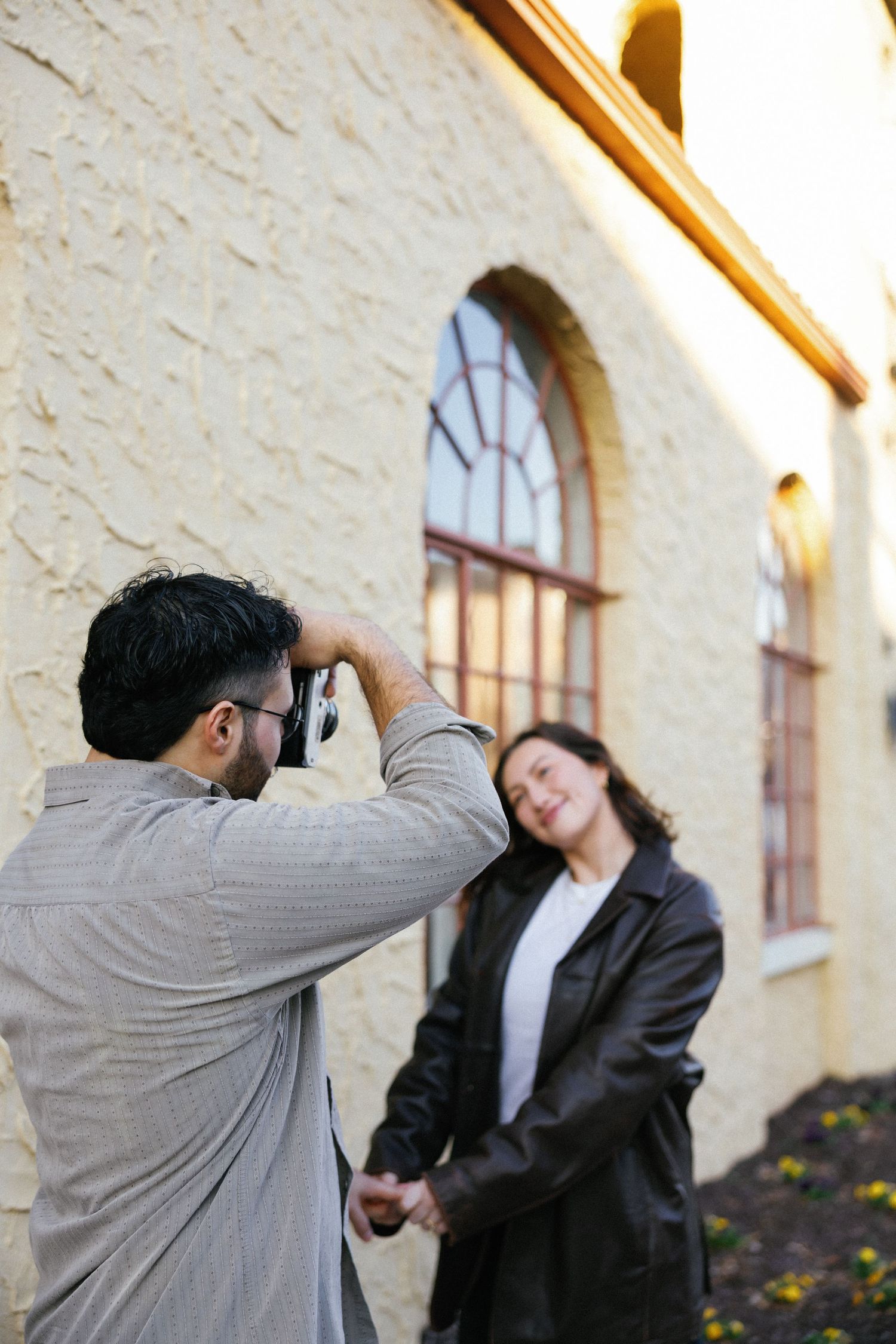 A photographer captures a smiling woman in a black jacket posing against a textured cream building wall outdoors.