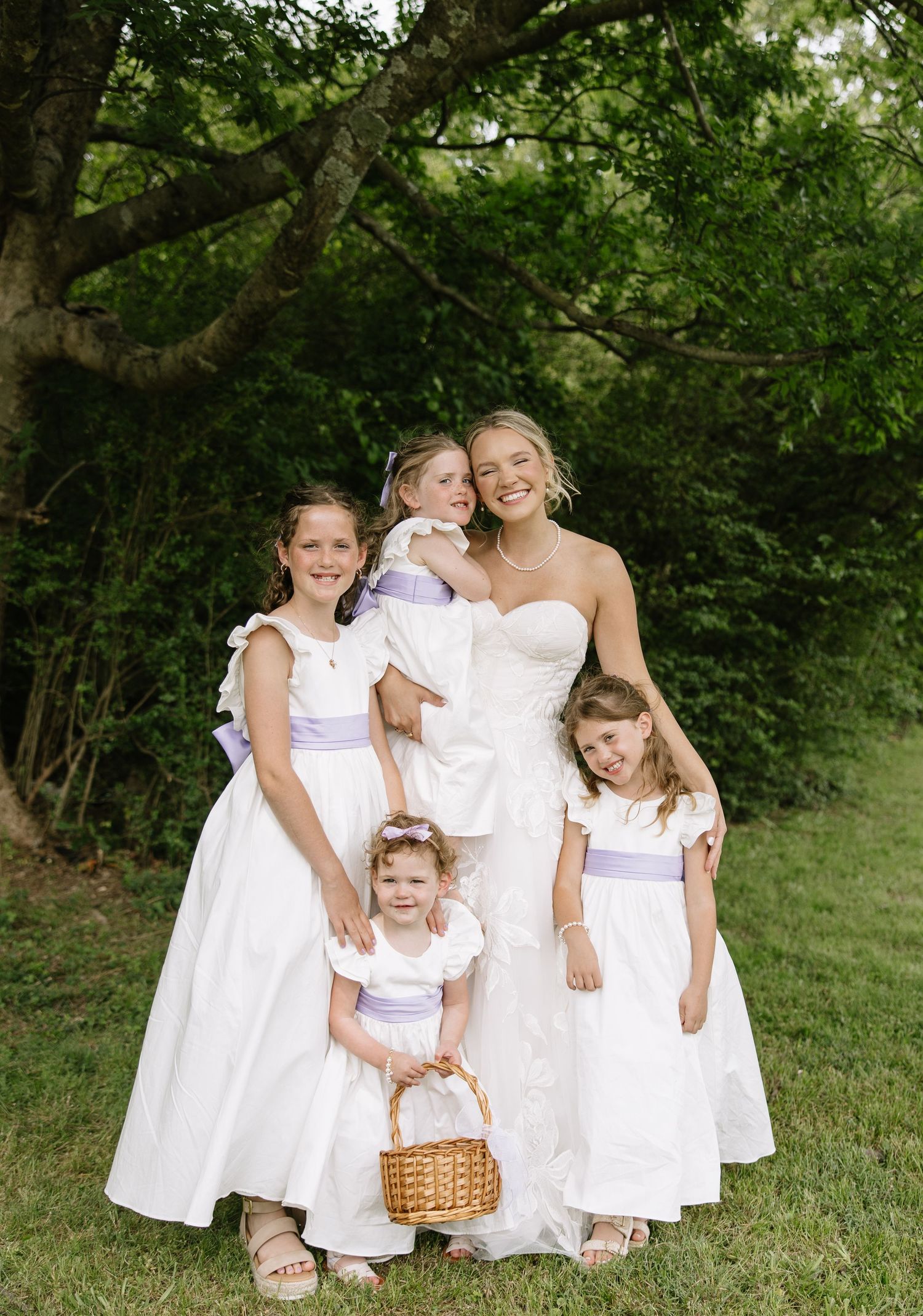 Bride in white strapless gown poses with four flower girls in white dresses with purple sashes outdoors.