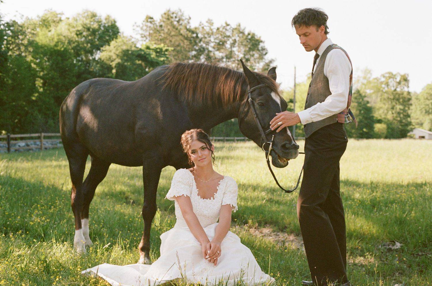 Bride in white dress sitting in grass beside groom holding a dark horse in a sunny field.