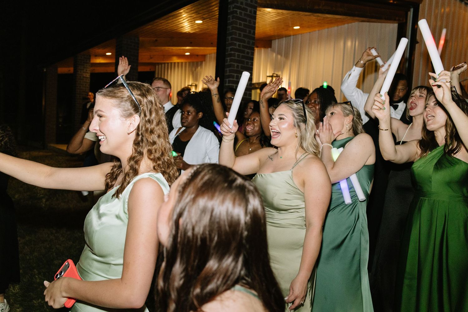 Bridesmaids in green dresses celebrate at outdoor wedding reception waving glow sticks with joyful energy.