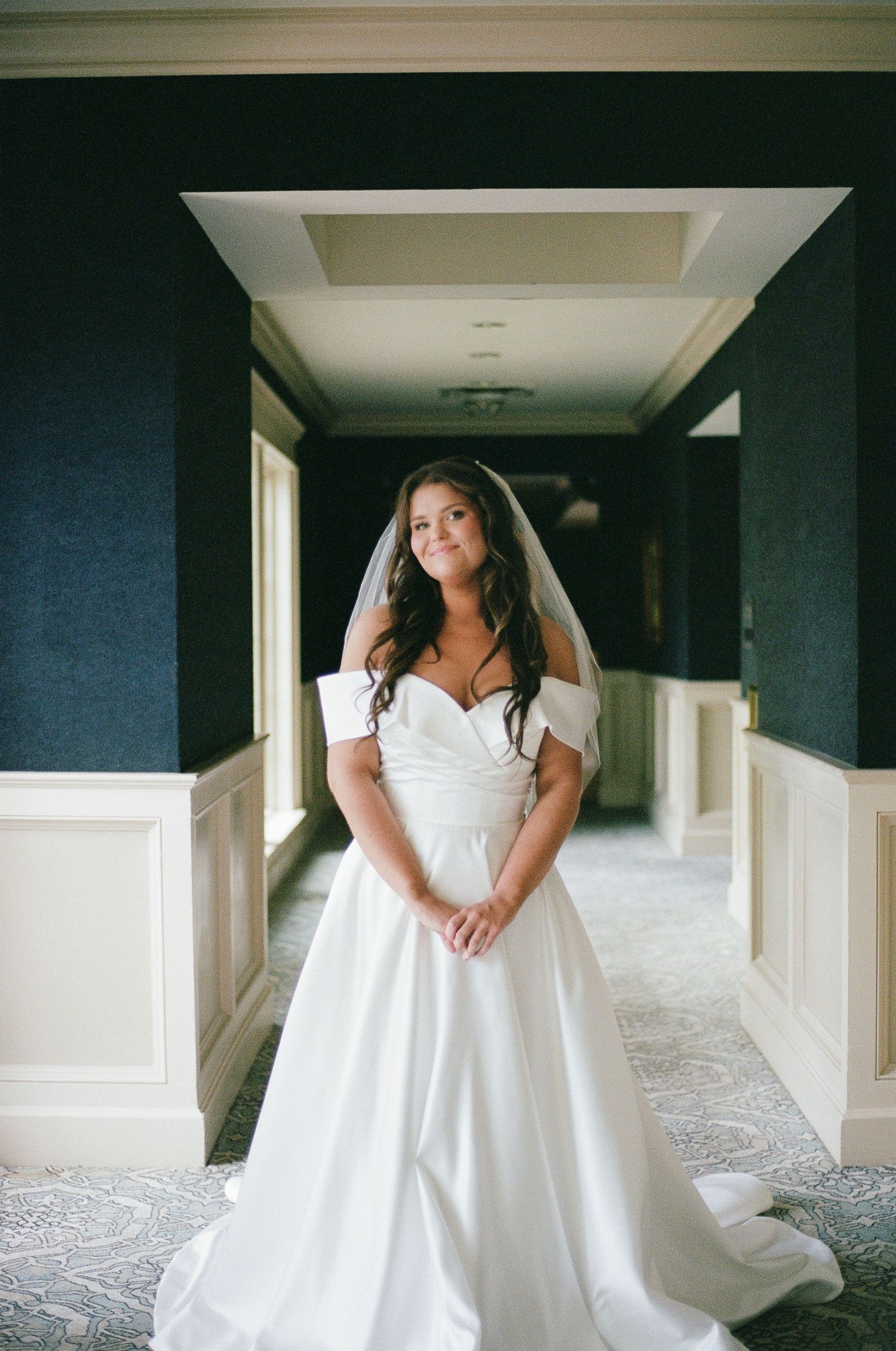 Bride in off-shoulder white ball gown with veil stands elegantly in a hotel corridor on her wedding day.