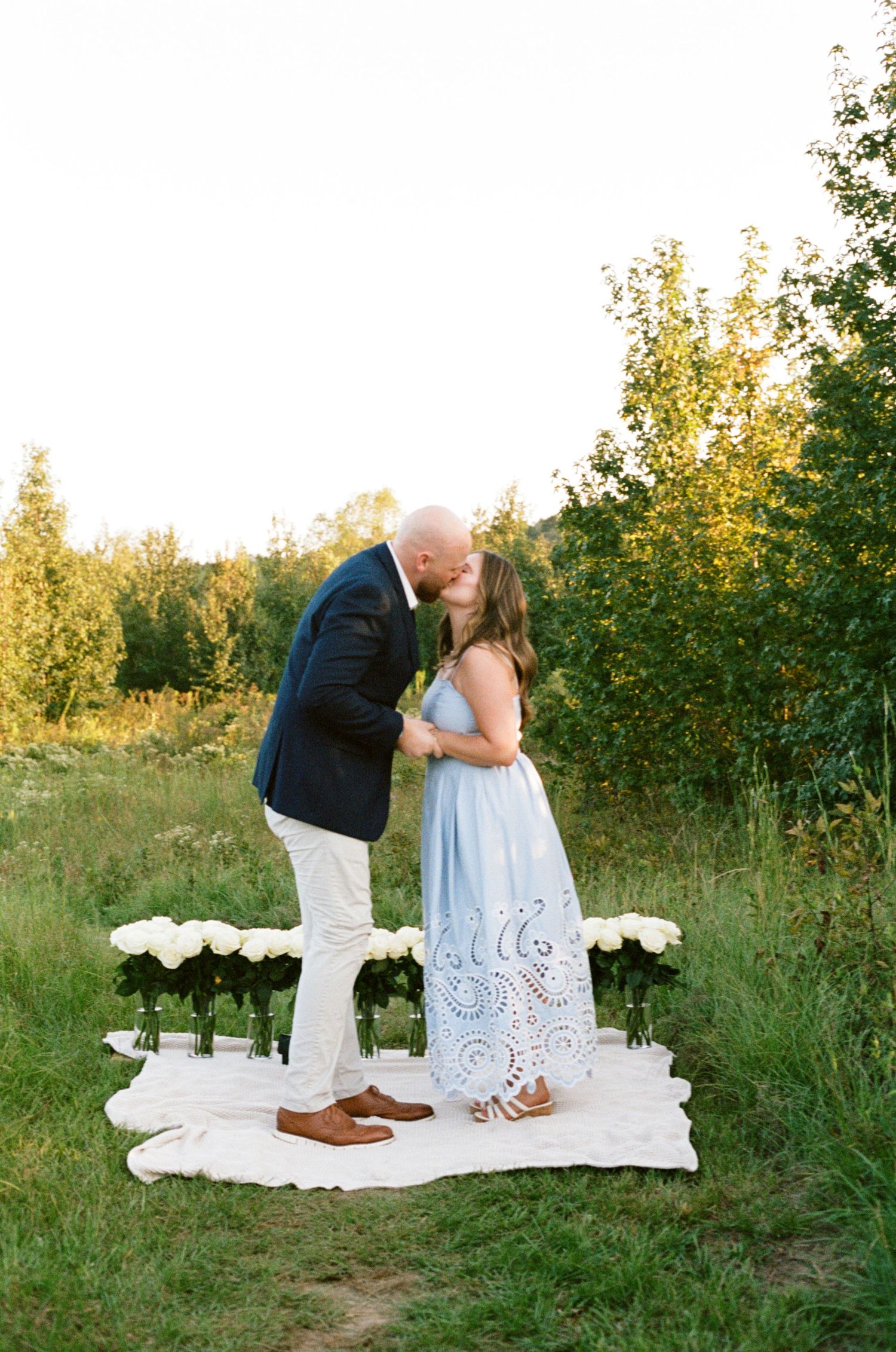 Couple kissing outdoors on white blanket with roses during golden hour in lush green field.
