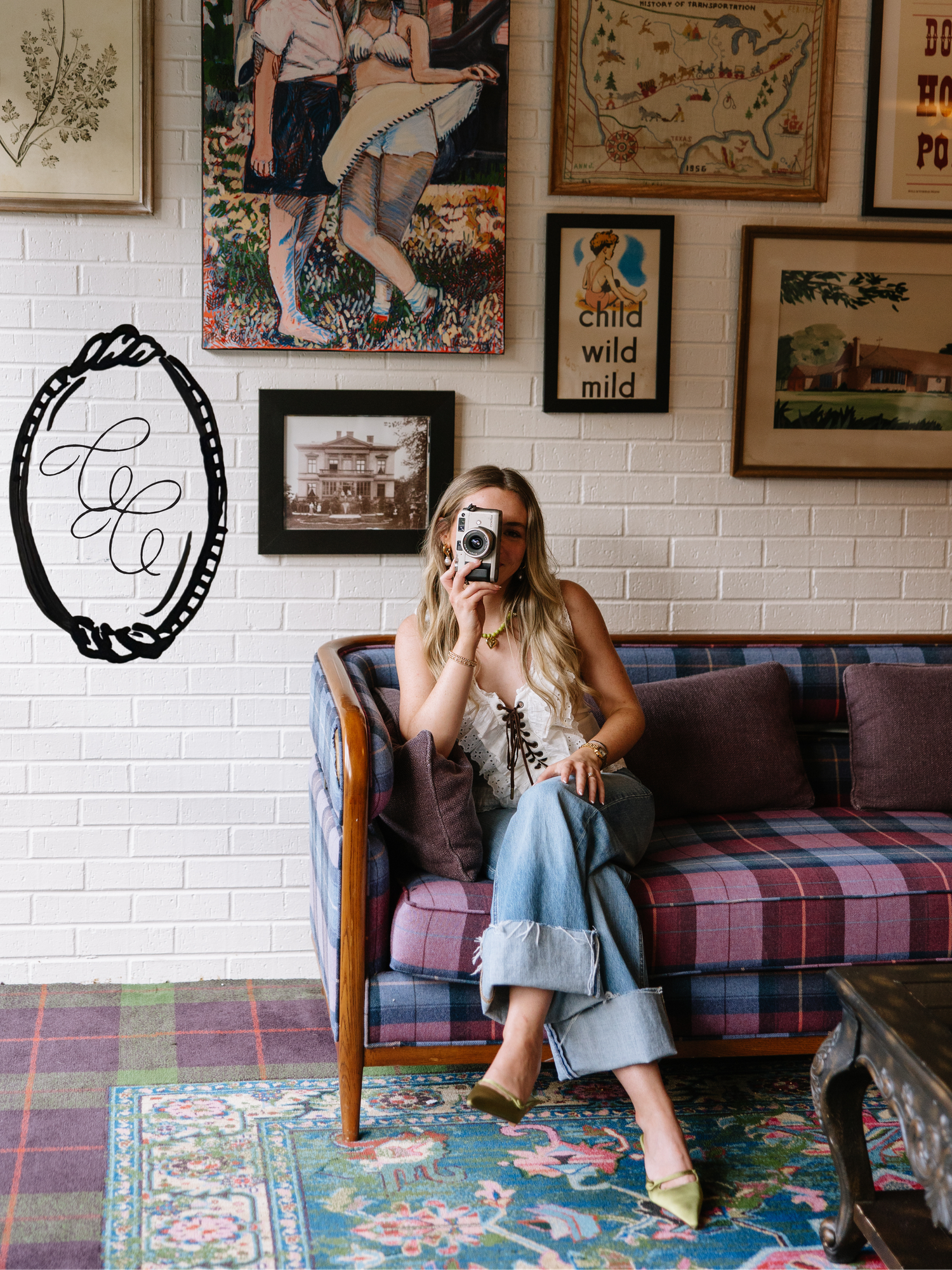 Woman sitting on plaid sofa holding camera, surrounded by eclectic wall art and vintage frames in a bohemian decorated room.