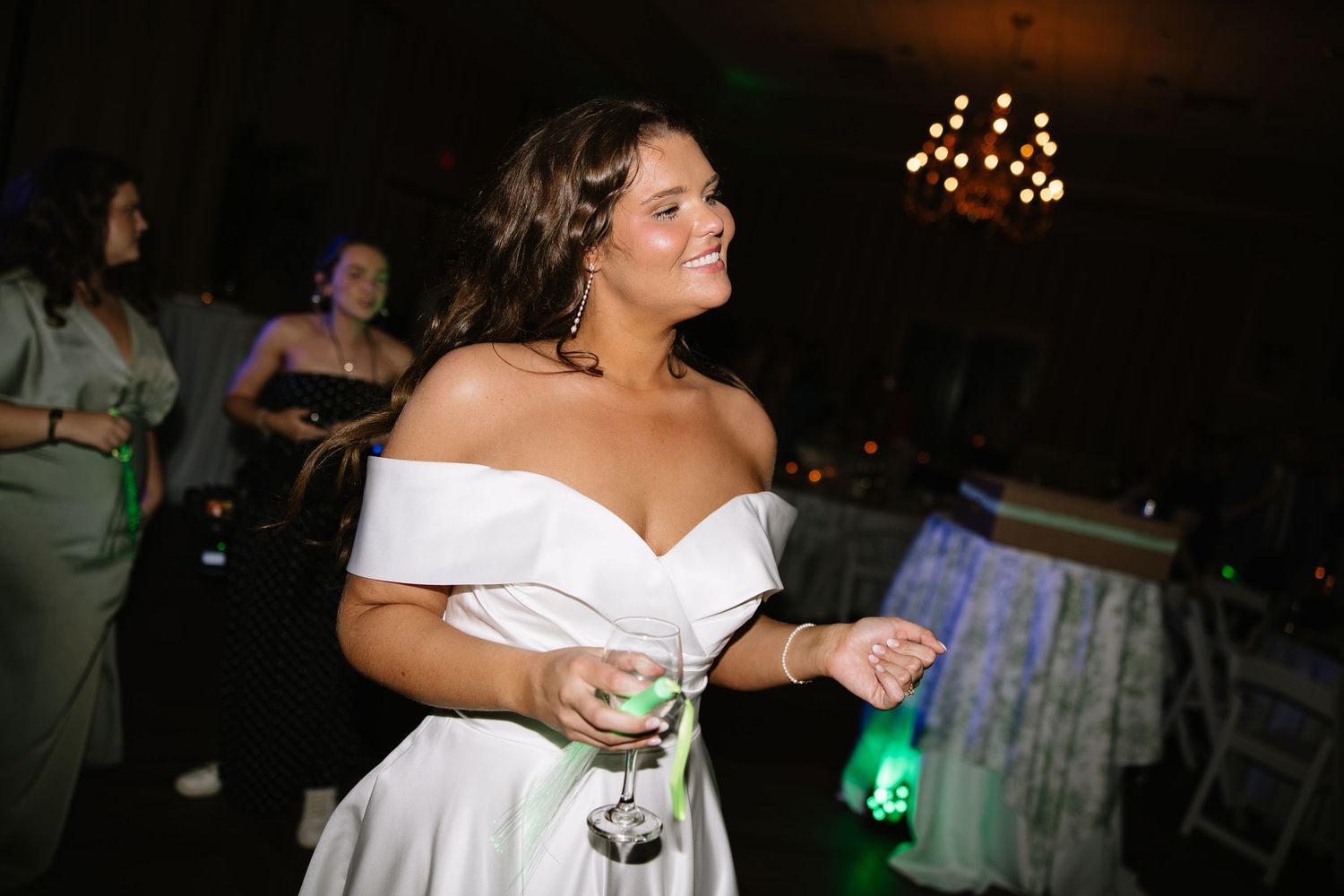 Bride in off-shoulder white dress holding a cocktail glass, smiling at a wedding reception.