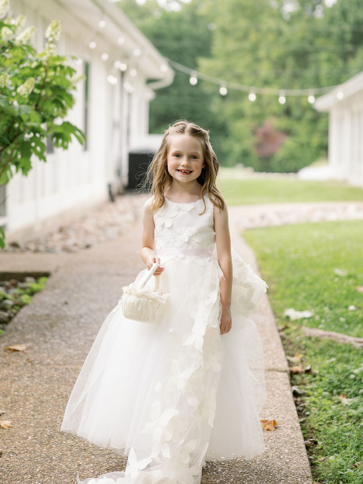 Young flower girl in white tulle gown carrying a floral basket walks along a garden pathway at a wedding.