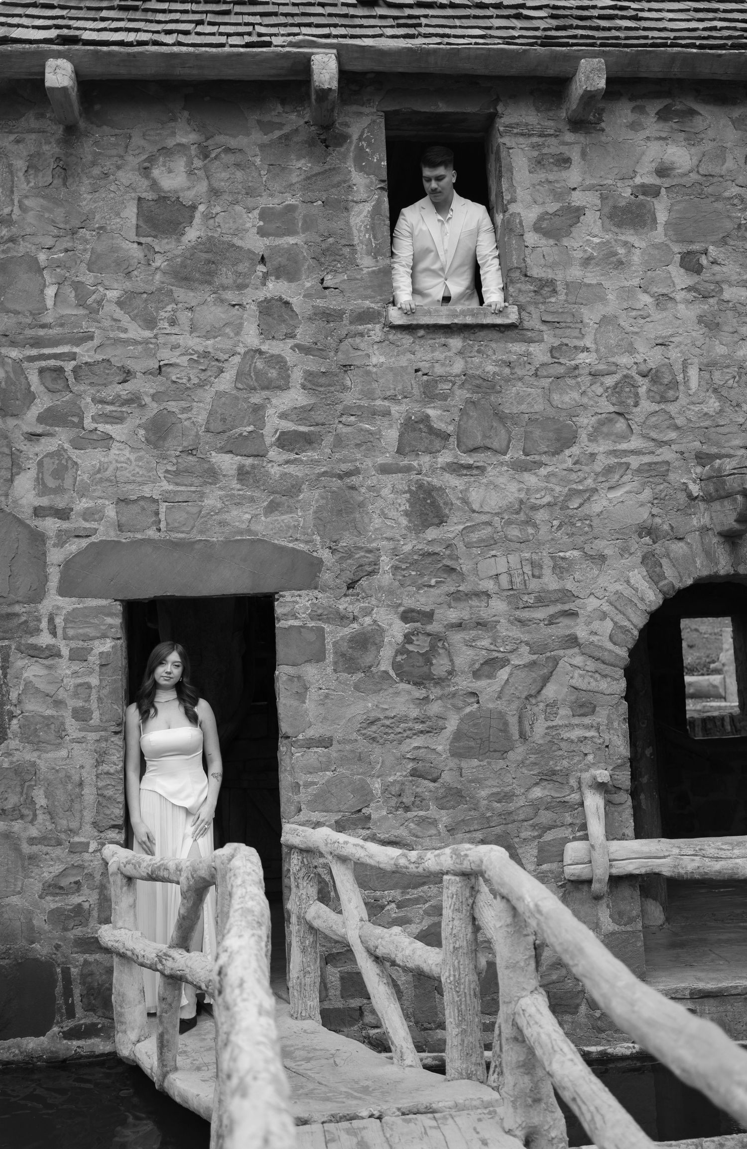 Black and white photo of couple posed separately at window and doorway of historic stone building.