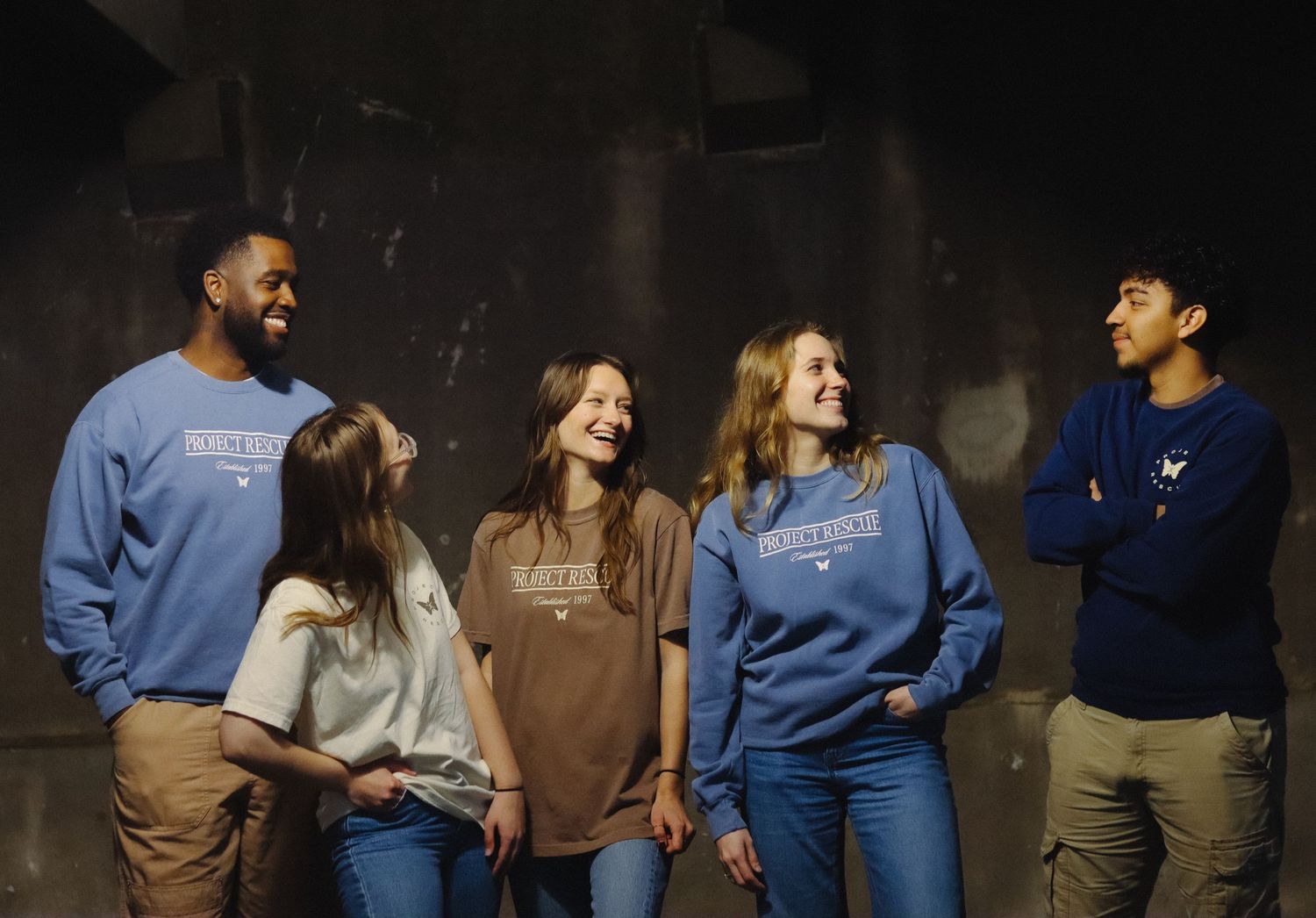 Five young adults laughing together wearing matching blue collegiate sweatshirts indoors.