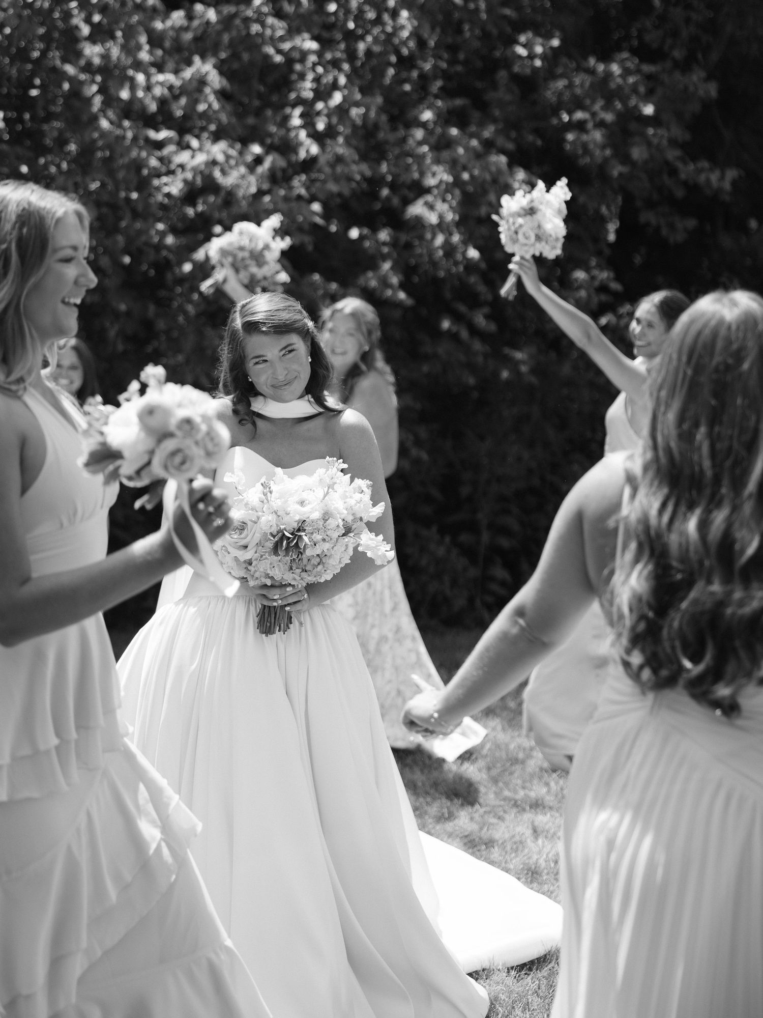Black and white photo of a smiling bride in a white gown surrounded by bridesmaids holding bouquets outdoors.