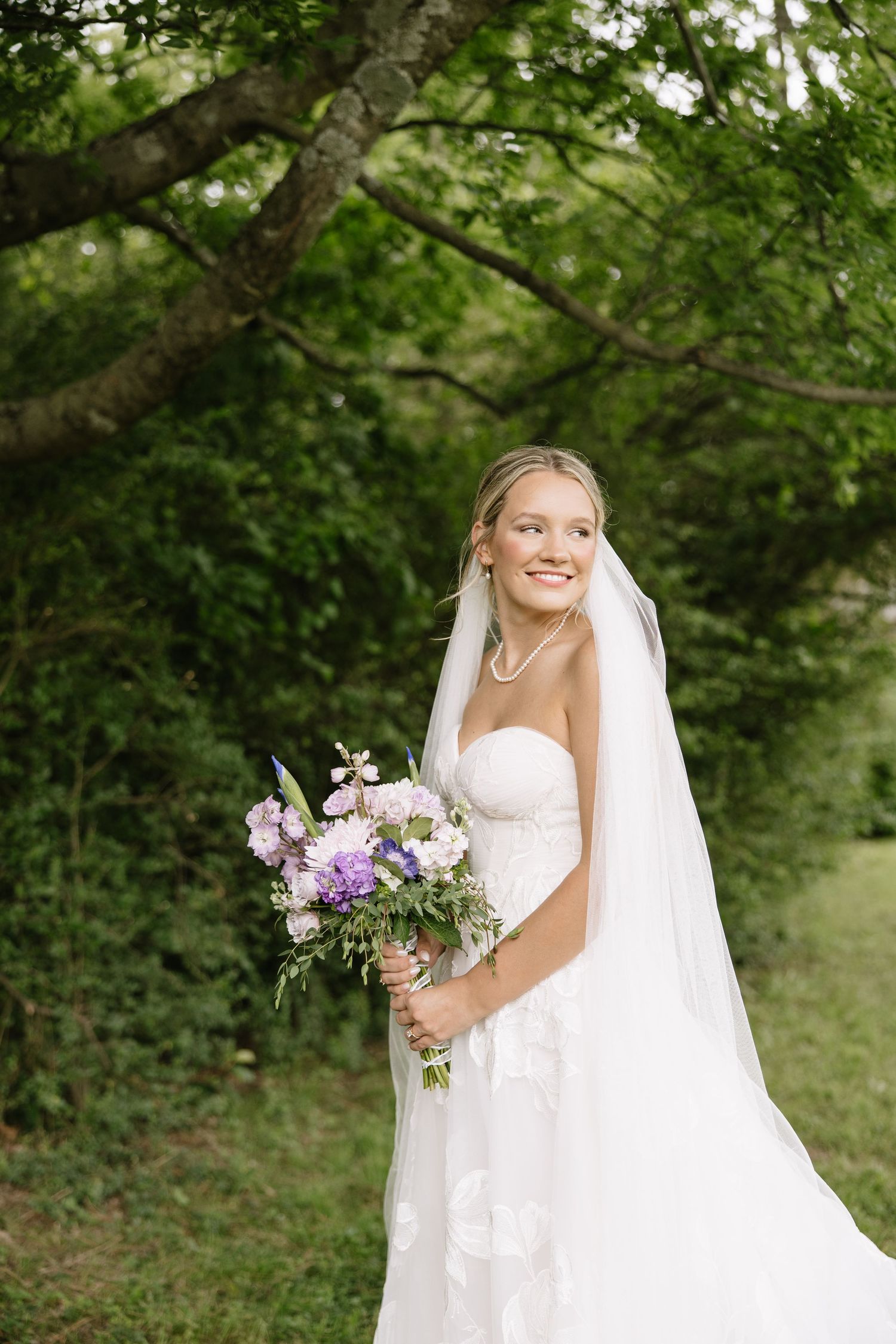 Smiling bride in strapless white gown and veil holds purple bouquet outdoors under lush green trees.