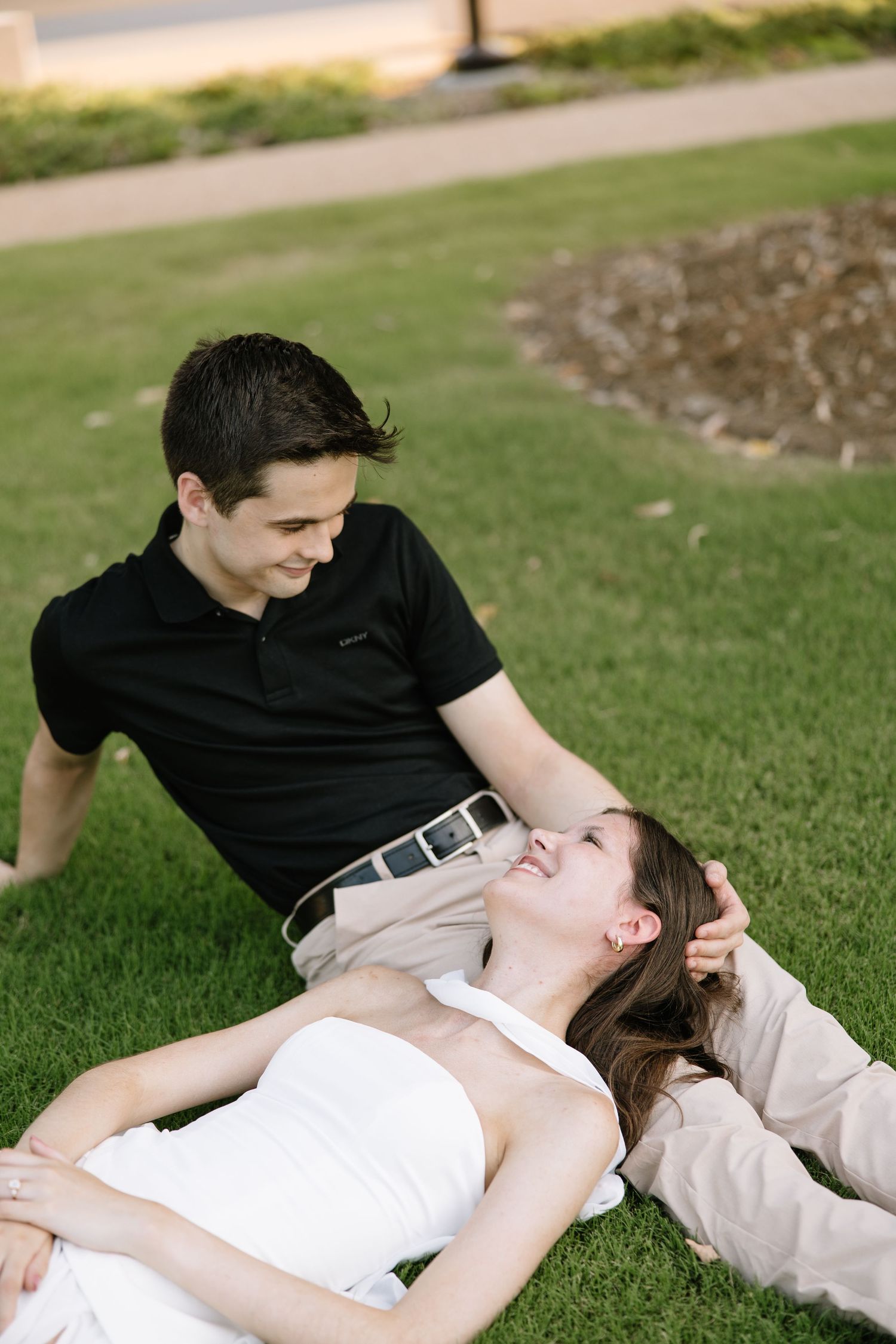 Couple relaxing on green grass, woman in white dress lying with head in mans lap, both smiling at each other.