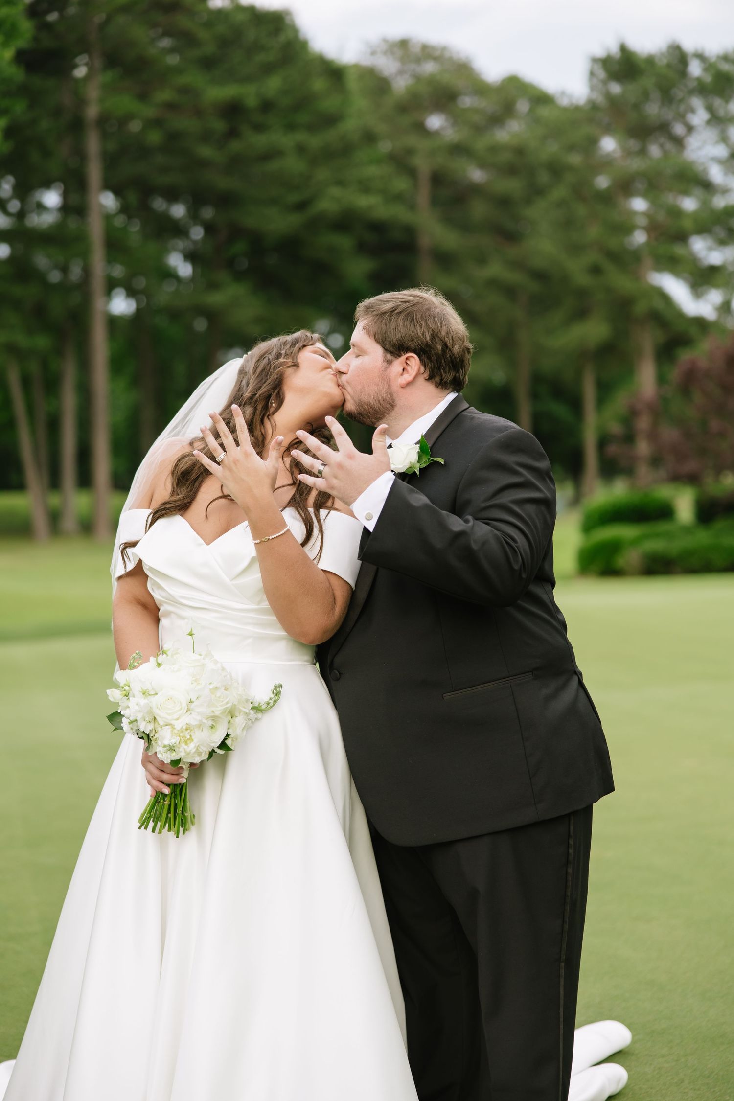 Bride and groom share a kiss outdoors on a golf course, bride holding white floral bouquet and showing rings.