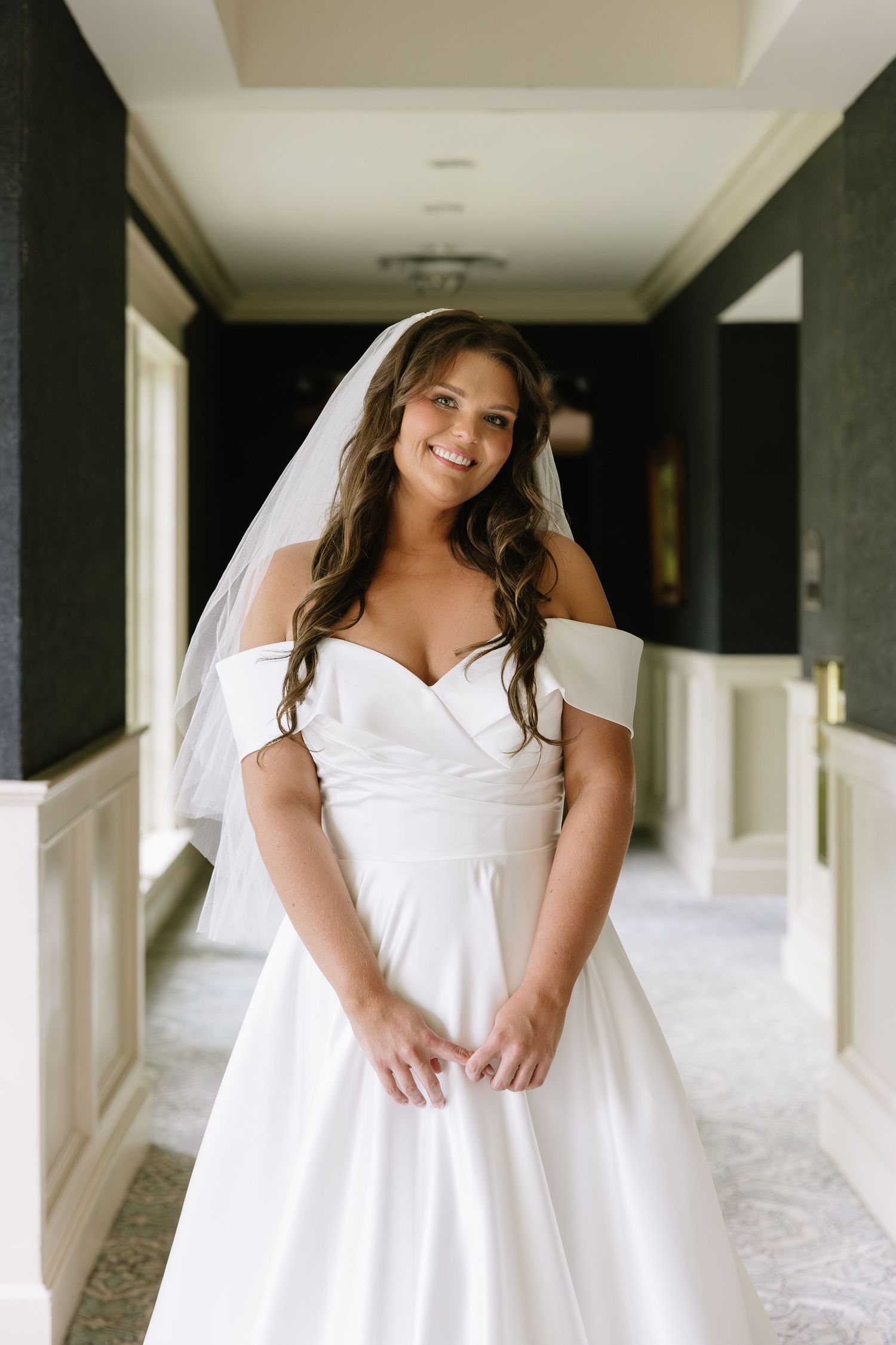 Smiling bride in off-shoulder white satin gown and veil poses in elegant hallway on wedding day.