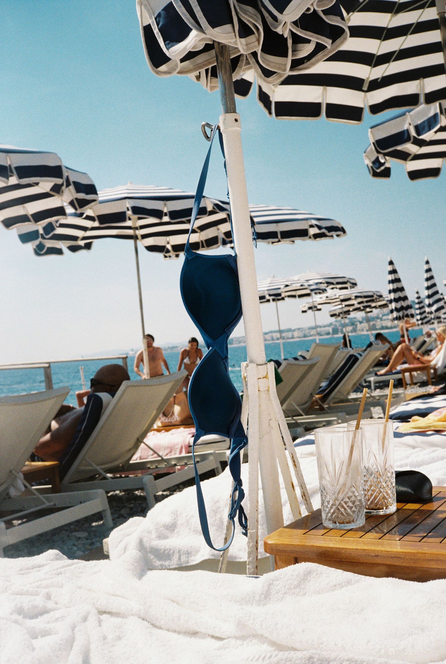 Blue bikini top hanging on a pole surrounded by striped beach umbrellas and sunbathers on lounge chairs.