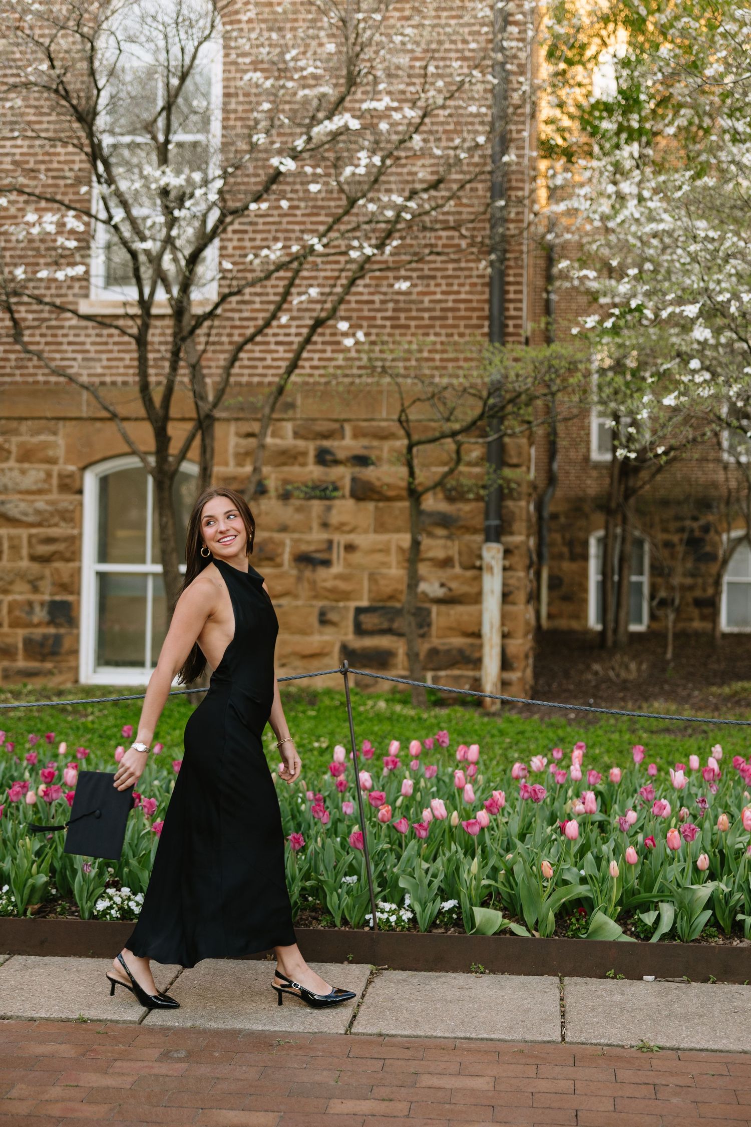 Woman in black halter dress walks past colorful tulip garden beside historic brick building.