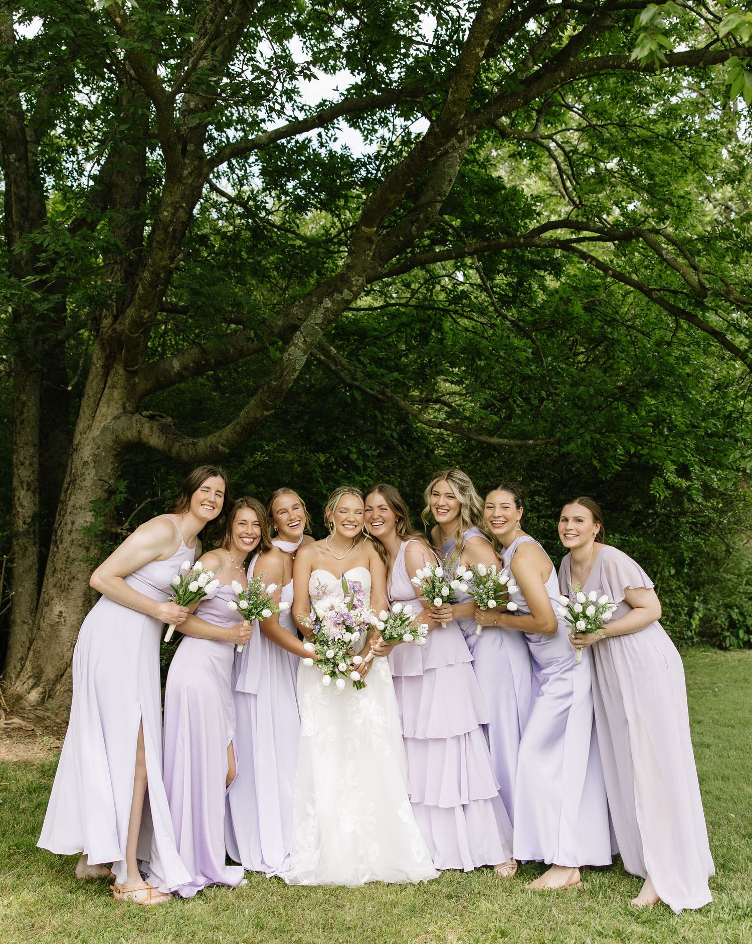 Bride in white gown poses with seven bridesmaids in lavender dresses holding floral bouquets under a large tree.
