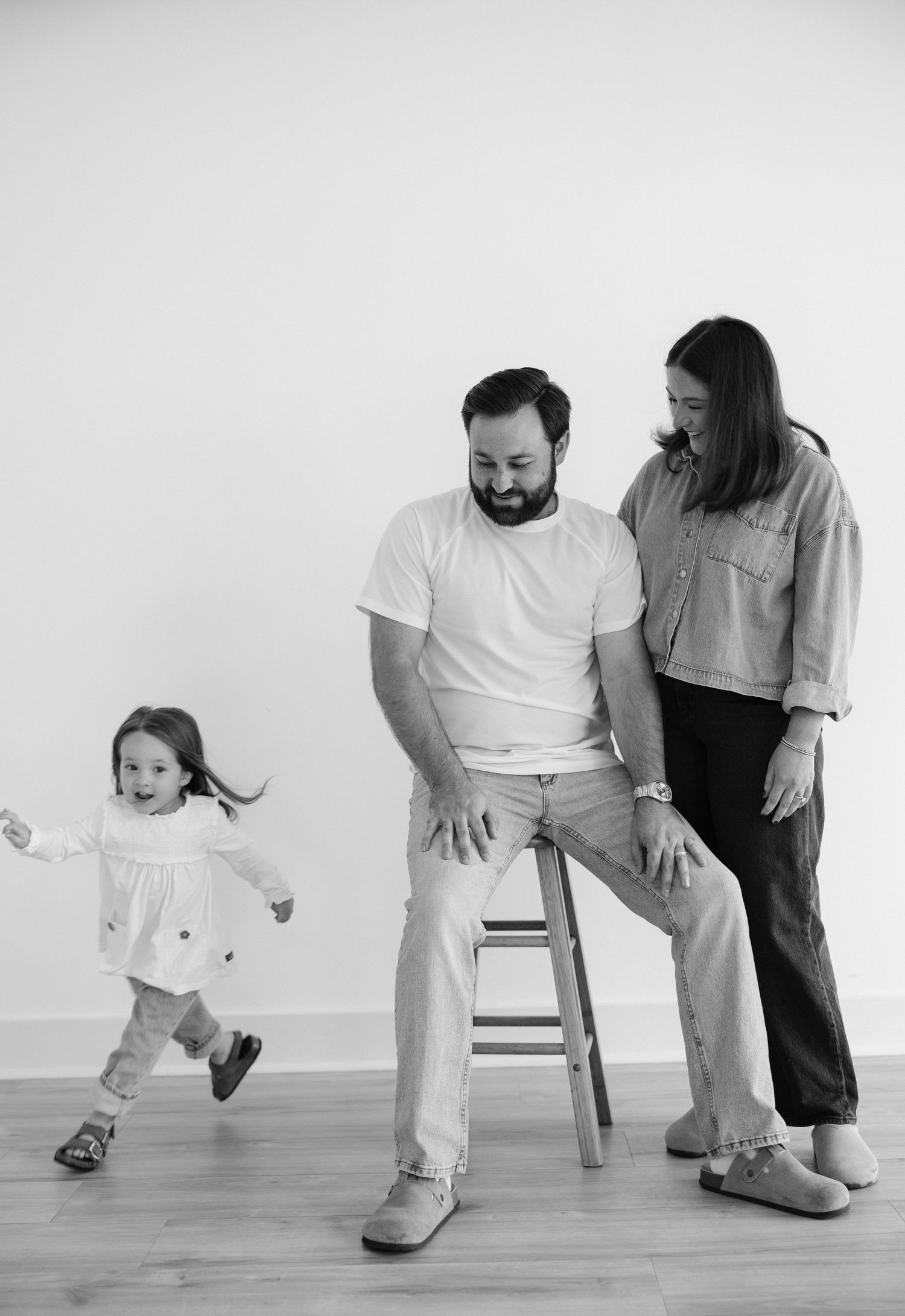 Black and white family photo of parents smiling at toddler girl running playfully in a bright studio setting.