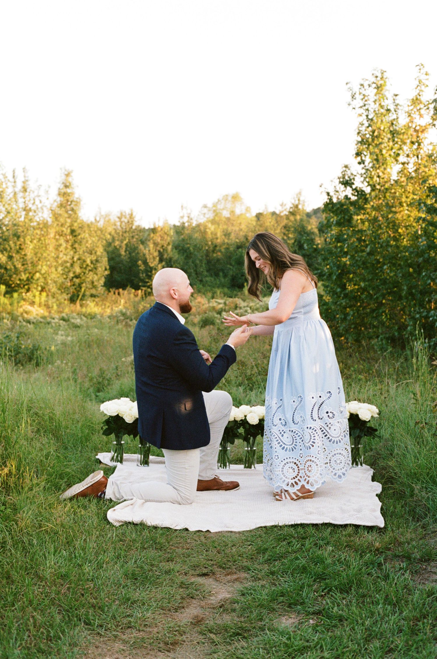 Man kneeling on white blanket proposing to woman in blue dress outdoors at golden hour with white roses.